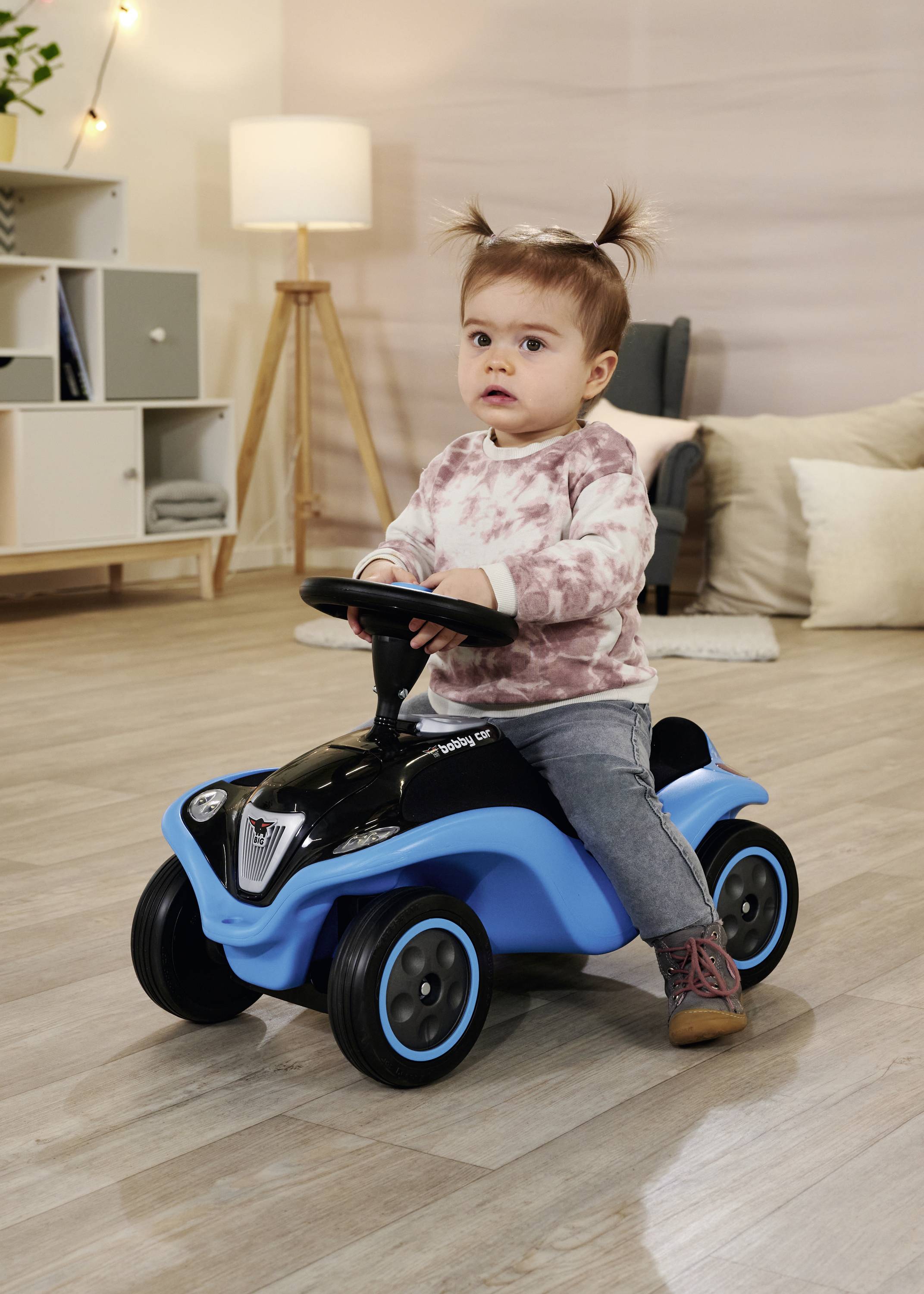 Ein Kleinkind mit zwei Zöpfen sitzt auf einem blauen Spielzeugauto in einem Wohnzimmer. Im Hintergrund sind Möbel und Dekorationen zu sehen.