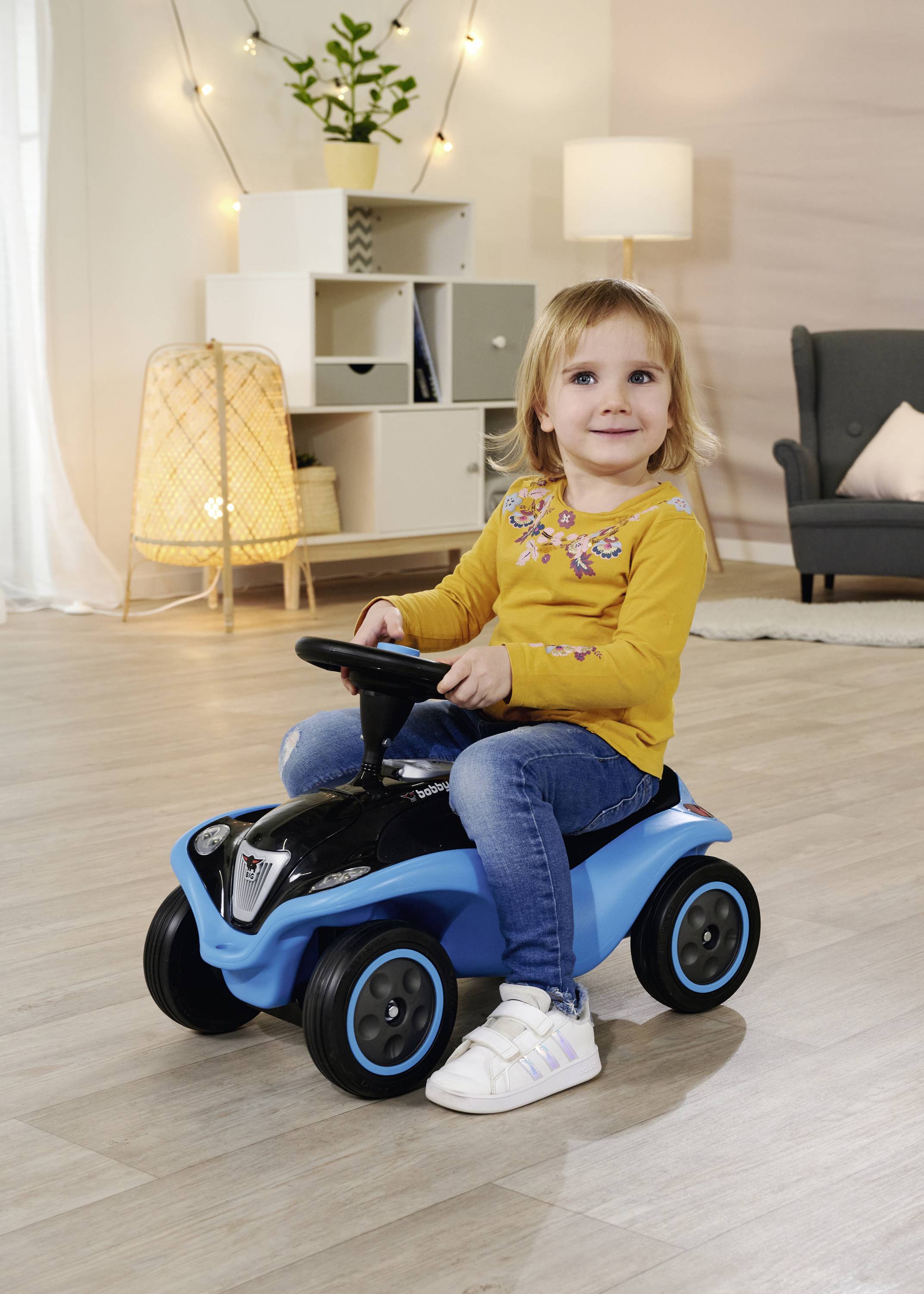 Ein Mädchen in einem gelben Pullover fährt in einem Wohnzimmer auf einem blauen Rutschauto. Im Hintergrund sind Möbel und Lampen.