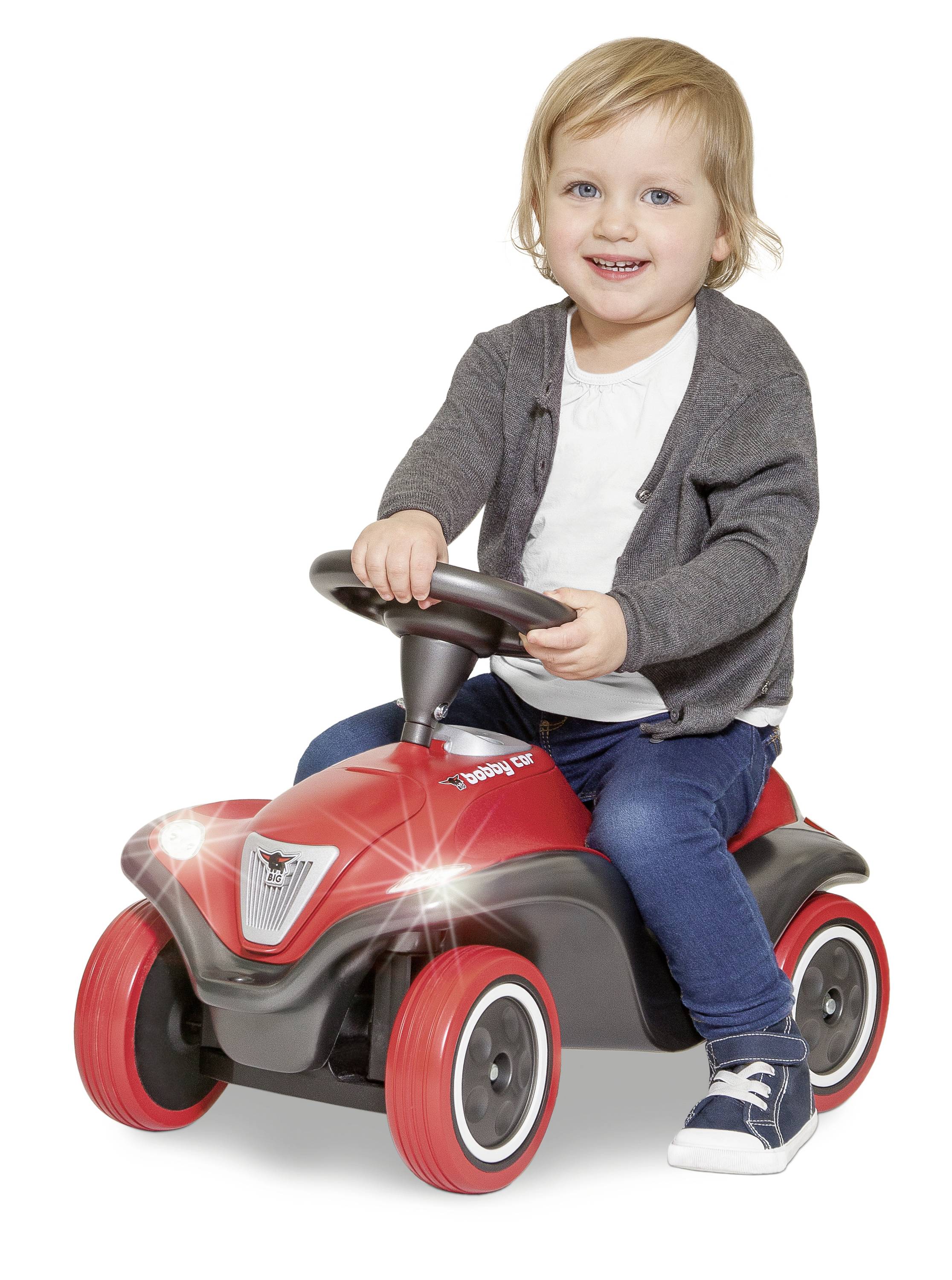 Ein Kind mit blonden Haaren sitzt lächelnd auf einem roten Spielzeugauto und trägt eine graue Jacke und blaue Jeans.
