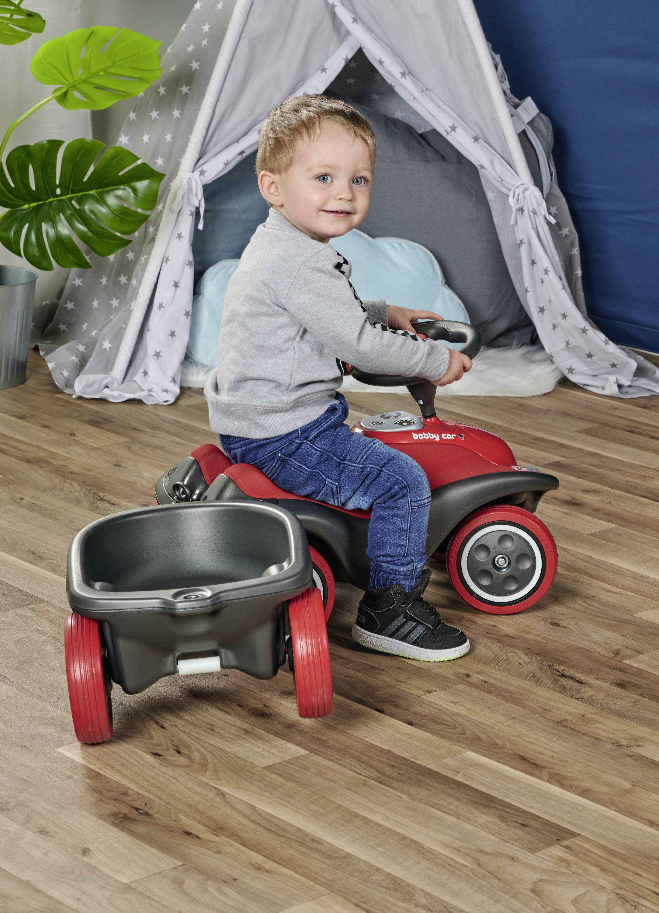 Ein kleiner Junge sitzt auf einem roten Spielzeugauto mit Anhänger in einem Kinderzimmer, lächelt und schaut zur Seite. Im Hintergrund ein Spielzelt.