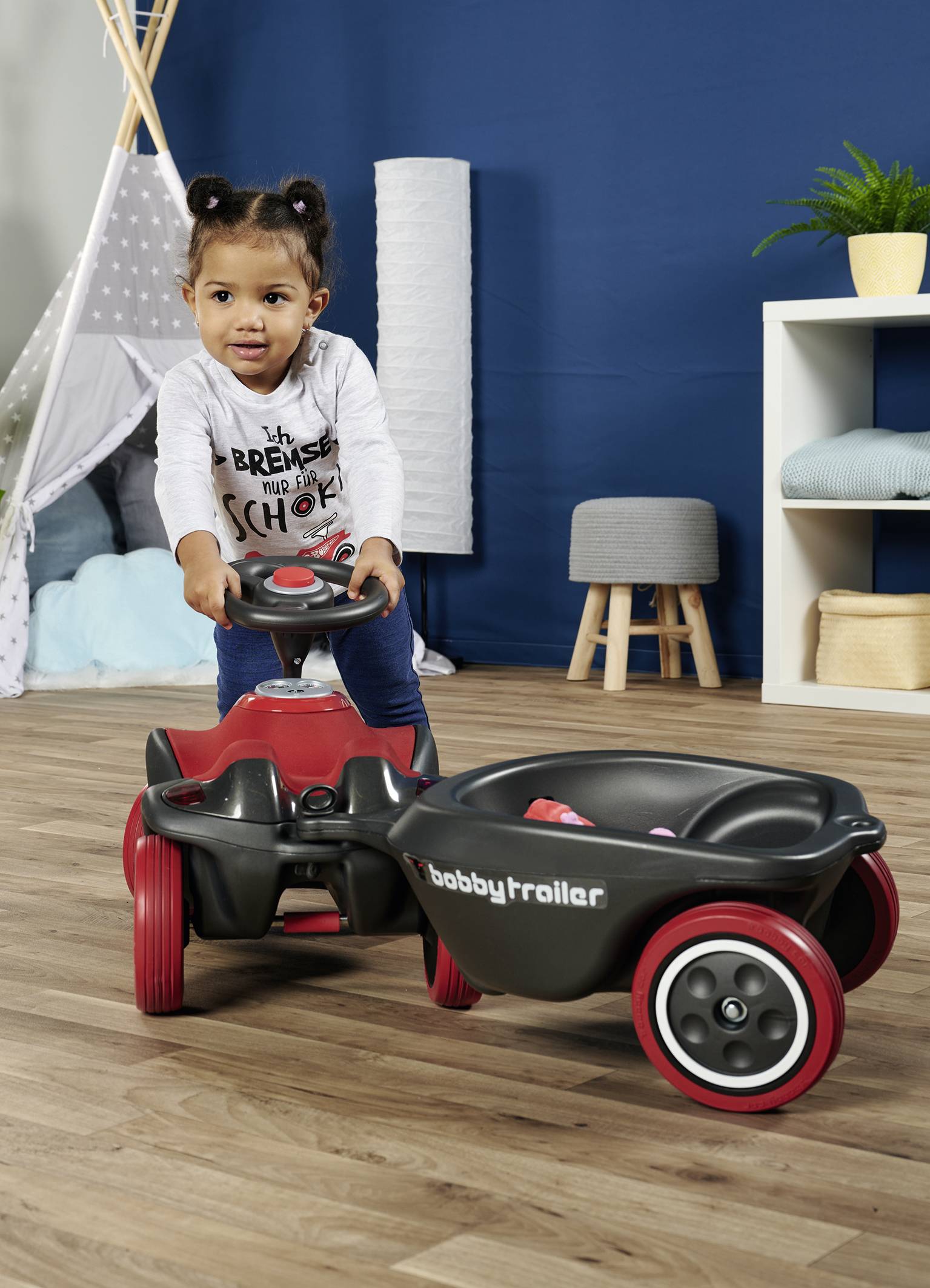 Ein Kind spielt mit einem roten und schwarzen Kinderrad im Wohnzimmer. Im Hintergrund sind ein Zelt und Regale zu sehen.
