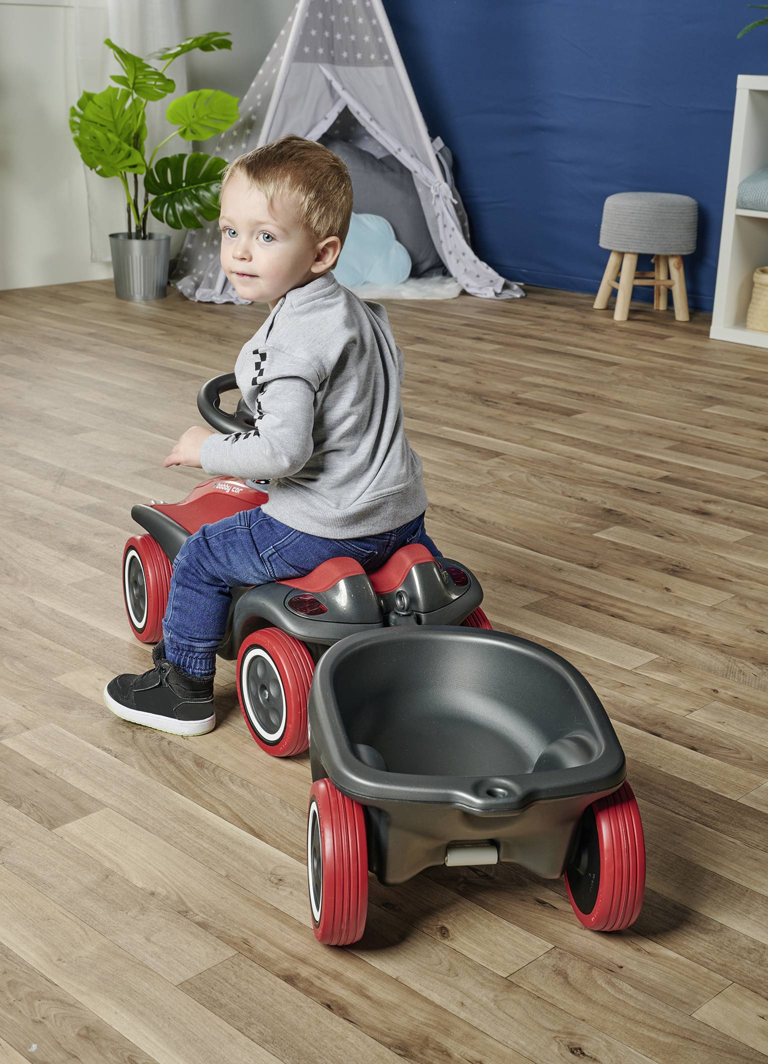 Ein Kind sitzt auf einem roten Spielzeugauto mit Anhänger in einem Zimmer. Im Hintergrund ist ein Zelt und eine Pflanze zu sehen.