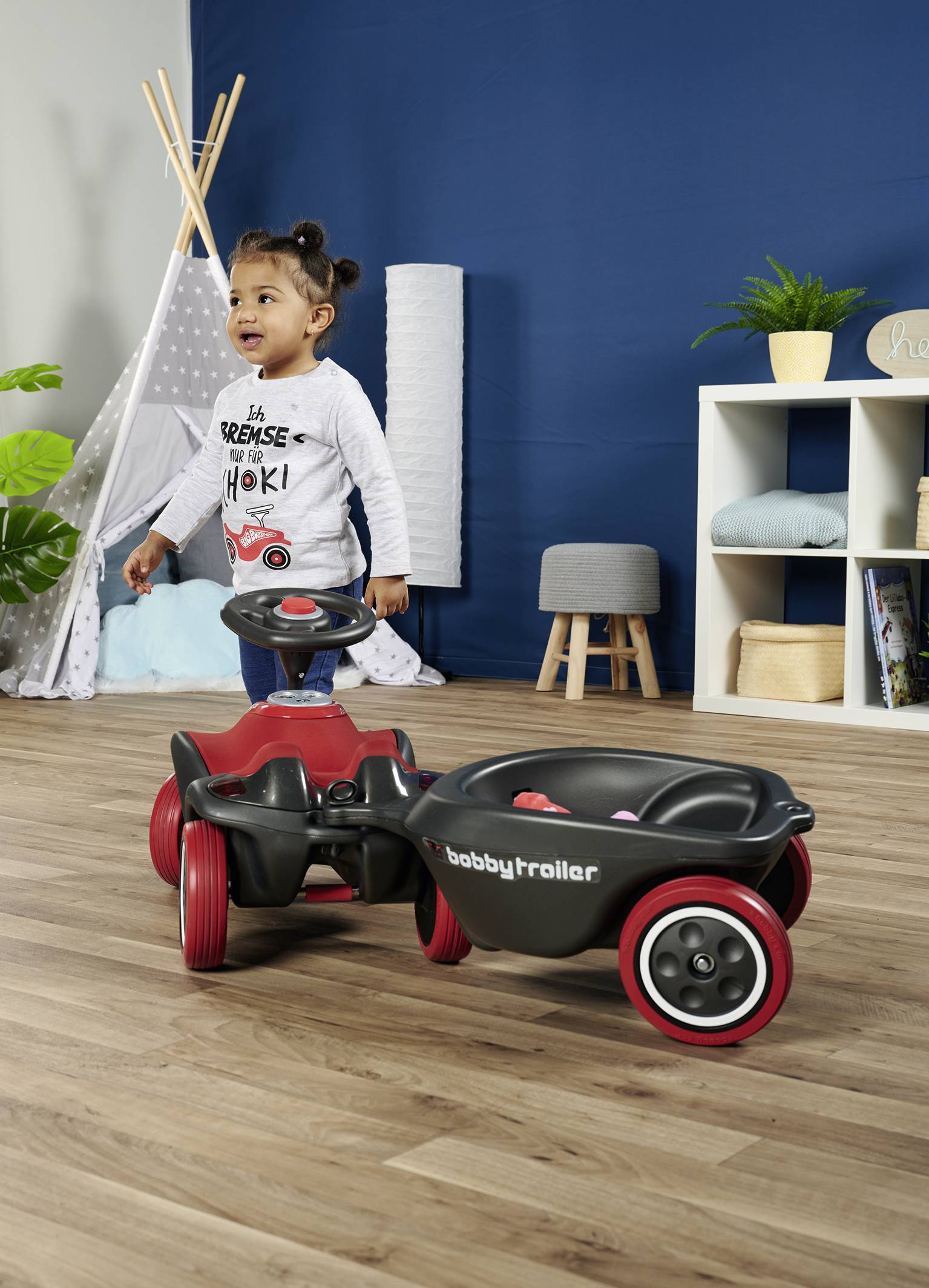 Ein Kind läuft fröhlich in einem Spielzimmer neben einem roten Kinderfahrzeug mit Anhänger. Im Hintergrund ein Regal und ein Zelt.