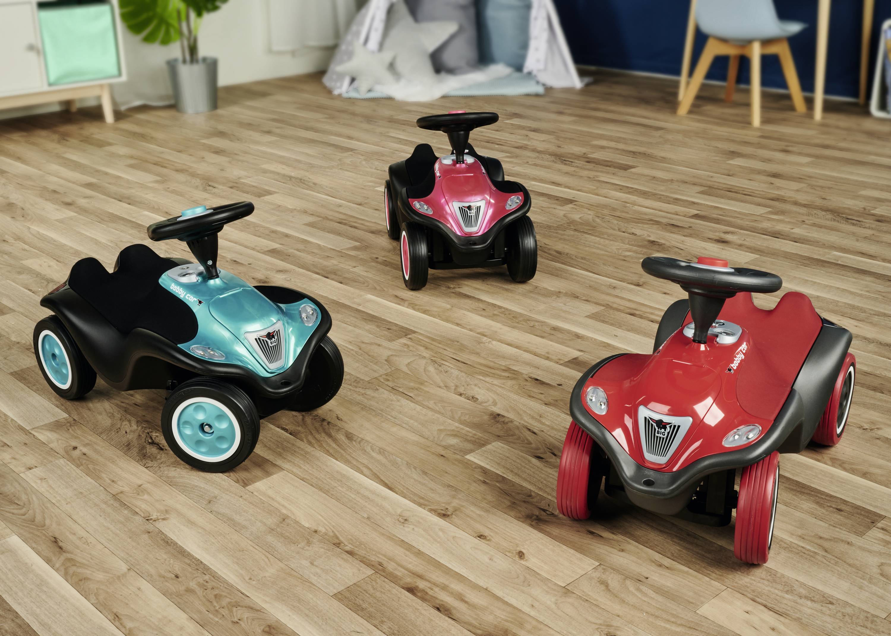 Drei Kinderfahrzeuge in einem hellen Raum, zwei rote und ein türkisfarbenes Auto, stehen auf einem Holzboden.