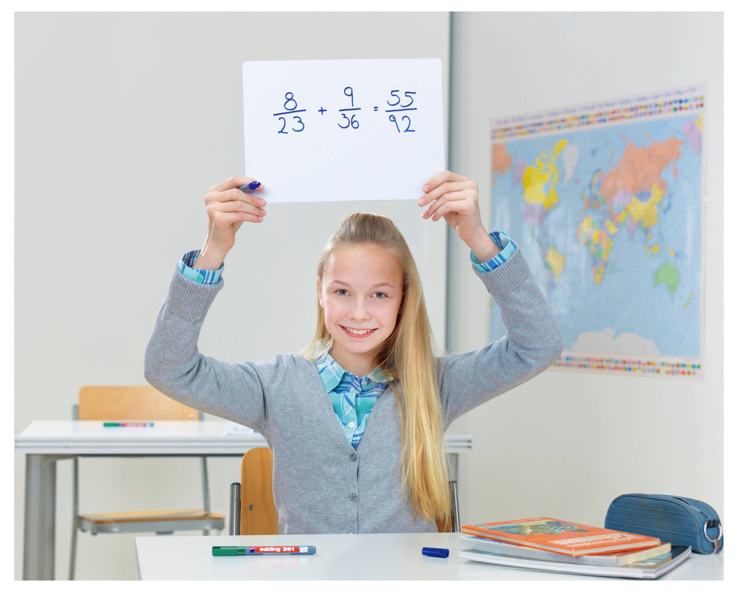 Lächelnder Schüler in einem Klassenzimmer hält ein Papier mit Bruchrechenaufgaben in die Höhe, mit einer Weltkarte und Schulmaterialien im Hintergrund.