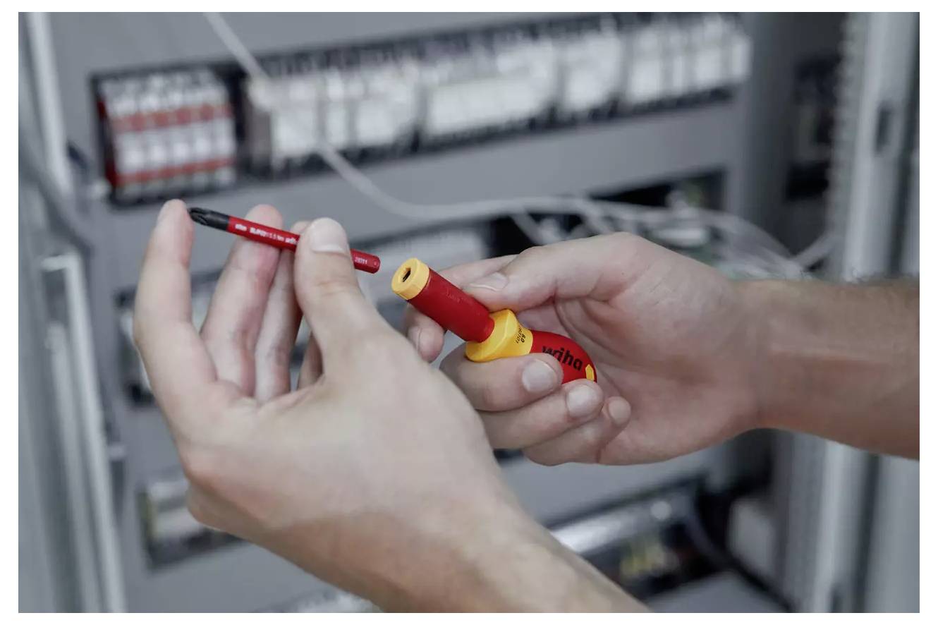 Eine Person hält einen rot und gelb isolierten Schraubendreher vor einem Elektroverteiler, was auf Arbeiten oder Wartungsarbeiten an der Verkabelung hinweist.