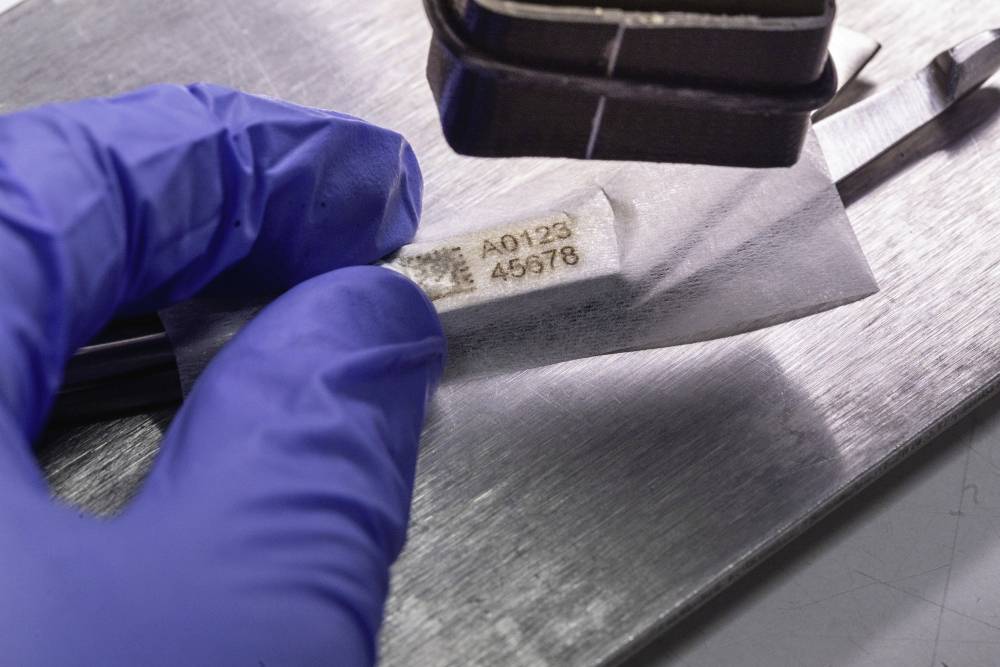 Eine Person hält eine Folie unter einem Werkzeug auf einem Metalltisch. Auf der Folie stehen die Nummern A0123 und 45678.