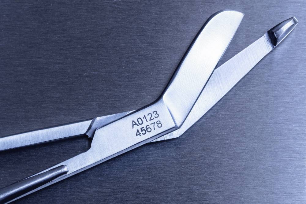 Metallisches chirurgisches Werkzeug mit Gravur 'A0123 45678', liegt geöffnet auf einem dunklen Hintergrund.