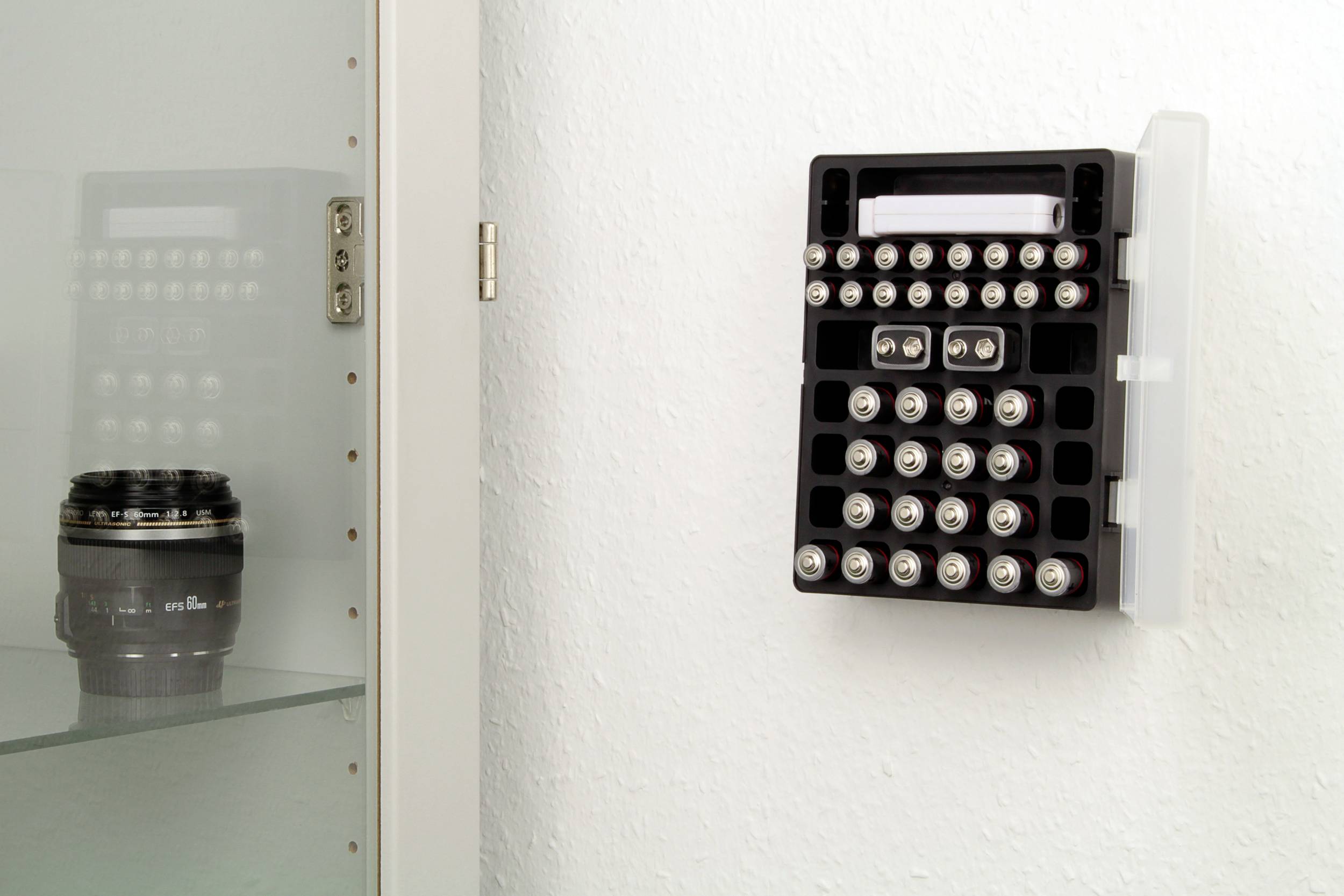 An der weißen Wand hängt ein schwarzer Batterienhalter mit verschiedenen Batterietypen. Links ist eine Kameraobjektiv in einem Glasregal zu sehen.