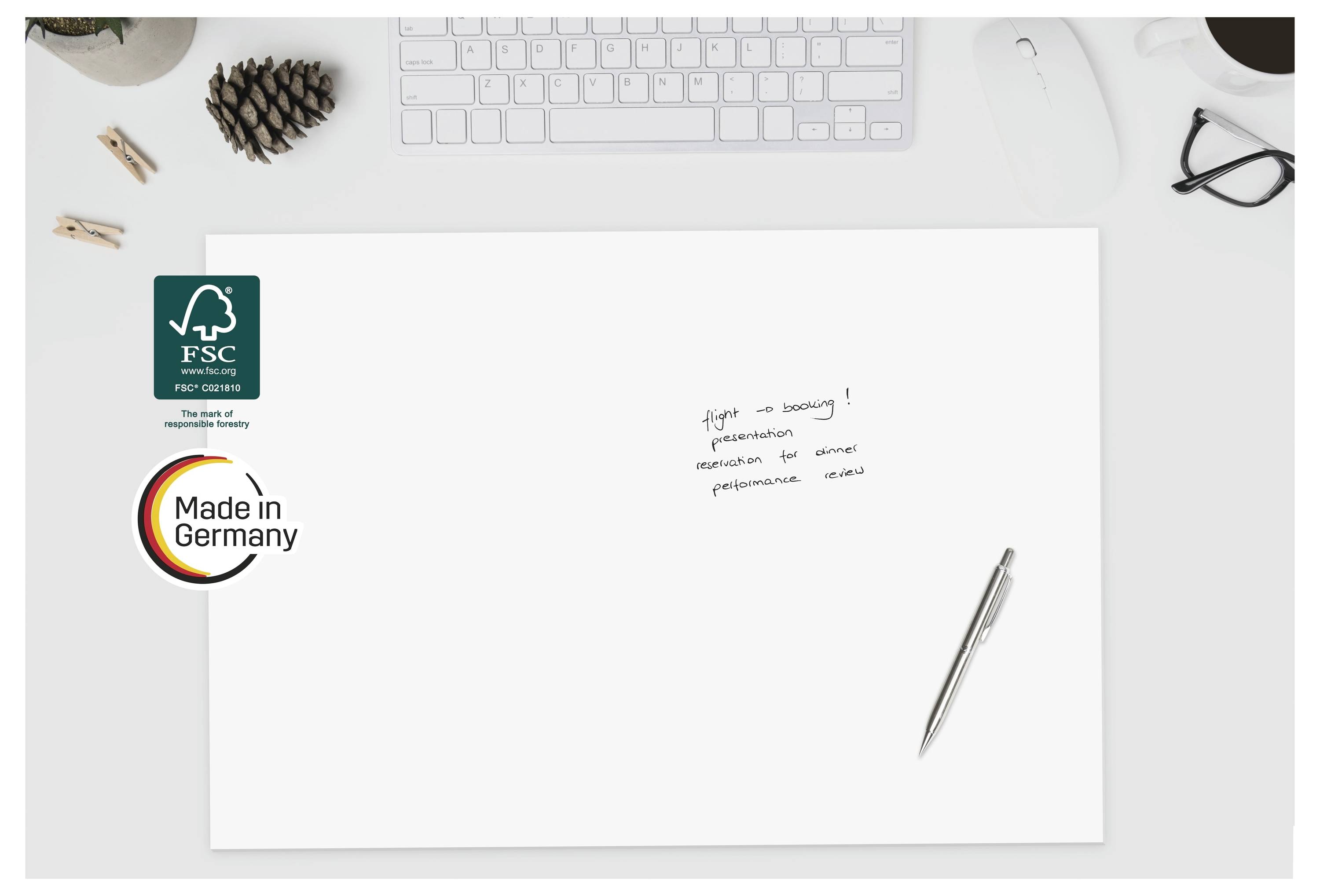 Leeres weißes Blatt auf einem Schreibtisch mit FSC- und Made in Germany-Logos, ein Stift, eine Tastatur, eine Brille und kleine handschriftliche Notizen auf der Seite.