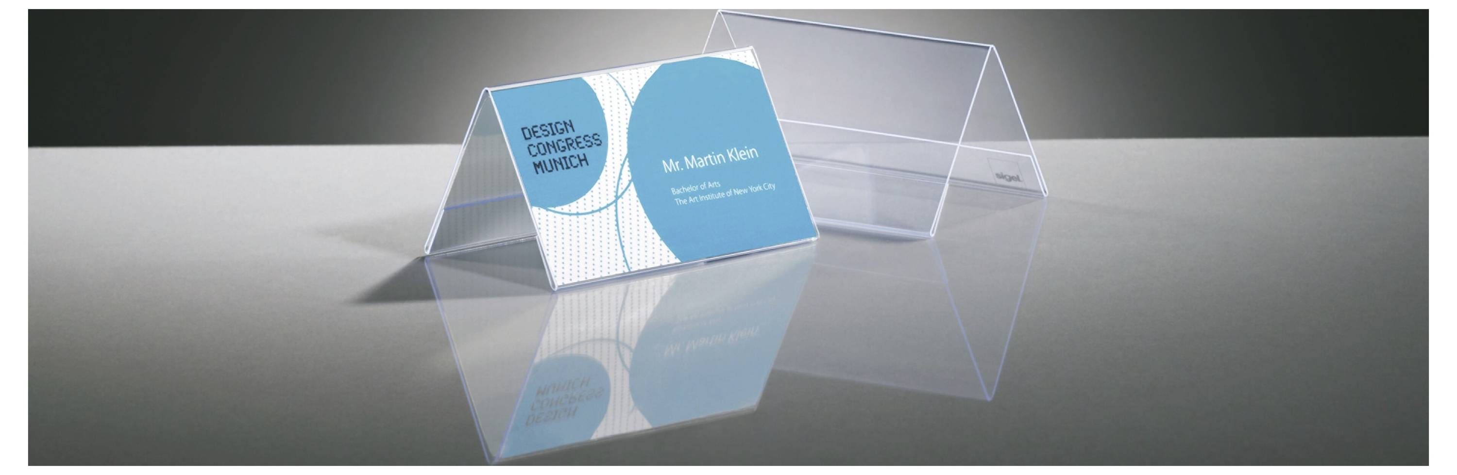 Klarer Acryl-Visitenkartenhalter, in dem eine blaue Visitenkarte von „Dazzle Custom Graphics" für Herrn Martin Klein auf einer reflektierenden grauen Oberfläche angezeigt wird.