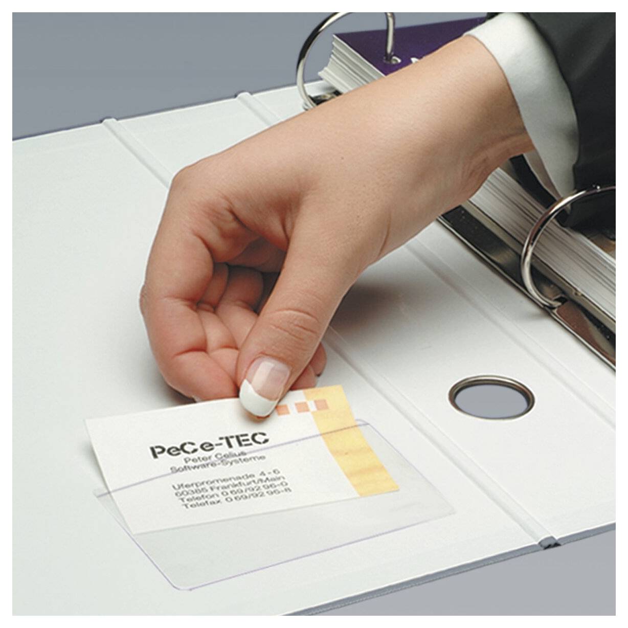 Hand legt eine Visitenkarte in eine durchsichtige Hülle in einem Ringordner ein, was auf Dokumentenorganisation oder Ablage hindeutet.