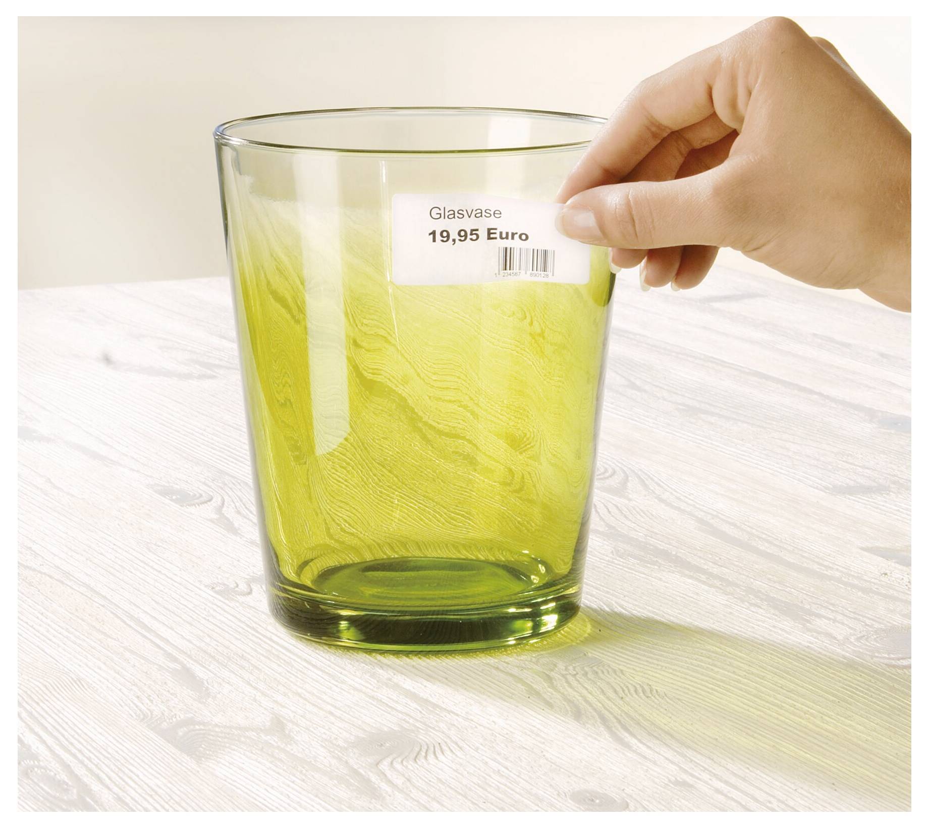 Hand, die ein Preisetikett mit der Aufschrift 'Glasvase 19,95 Euro' auf eine grüne Glasesvase auf einem hellen Holztisch legt.