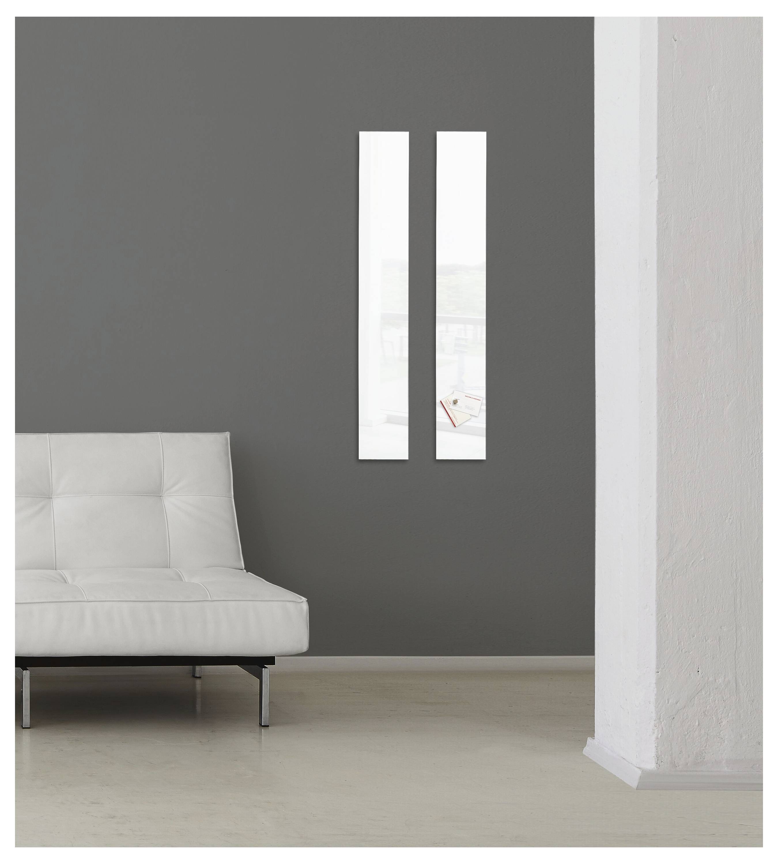 Minimalistische Inneneinrichtung mit einem weißen Sofa vor einer dunkelgrauen Wand, zwei schmalen vertikalen Fenstern und einer weißen Säule auf der rechten Seite.