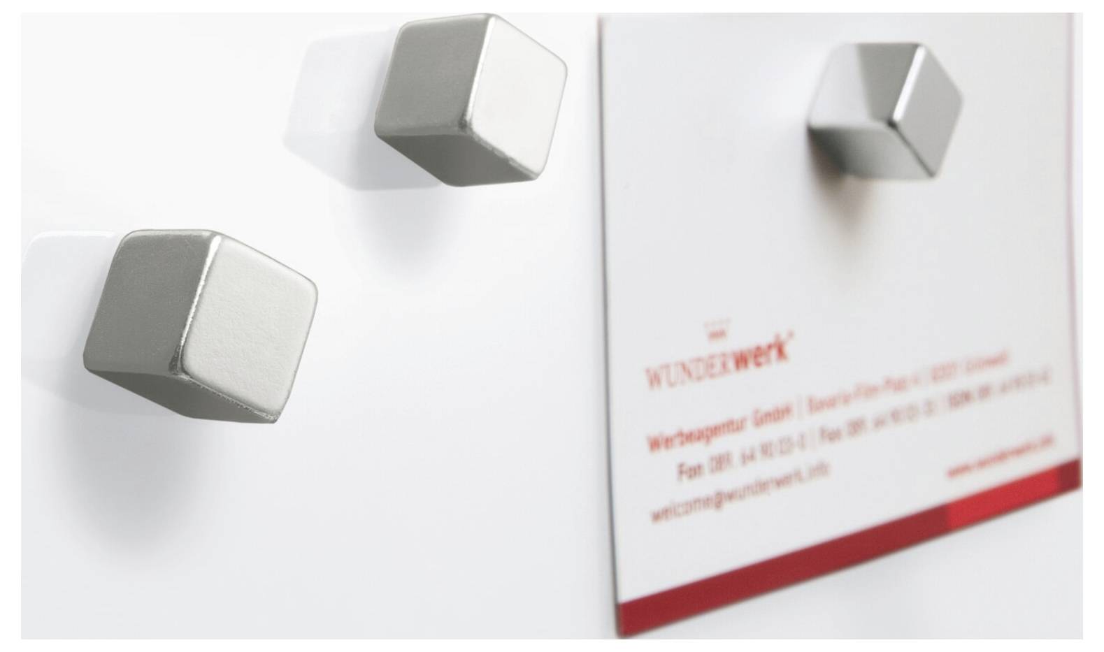 Nahaufnahme einer weißen Broschüre oder Karte mit drei kleinen silbernen Würfelmagneten, die ein unscharfes rotes "WUNDERWERK"-Text und einen roten Streifen zeigen