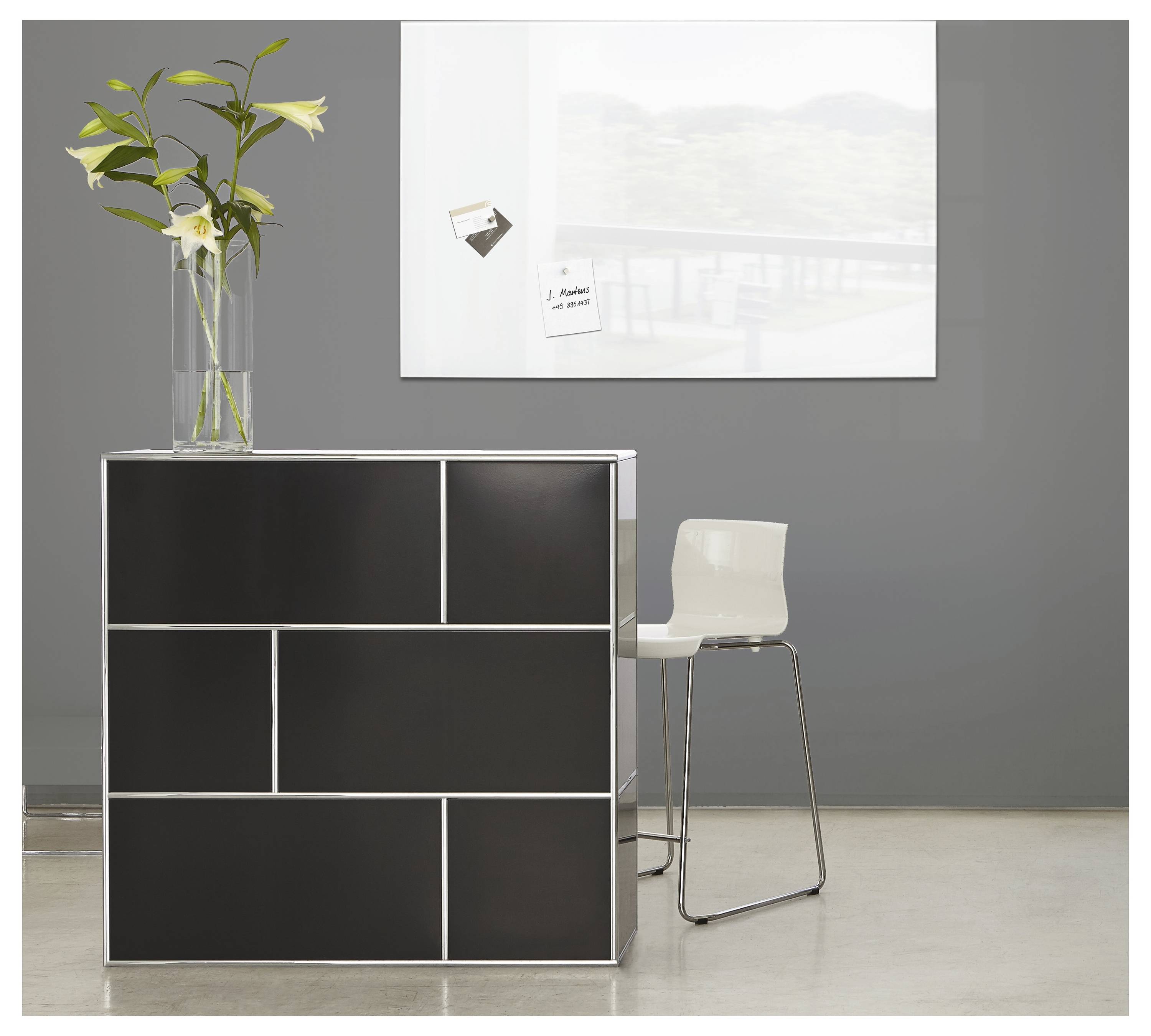 Modernes Lobby-Interieur mit schwarzem modularem Schrank, weißem Stuhl und Vase mit weißen Blumen vor einer grauen Wand mit Rahmenkunst.