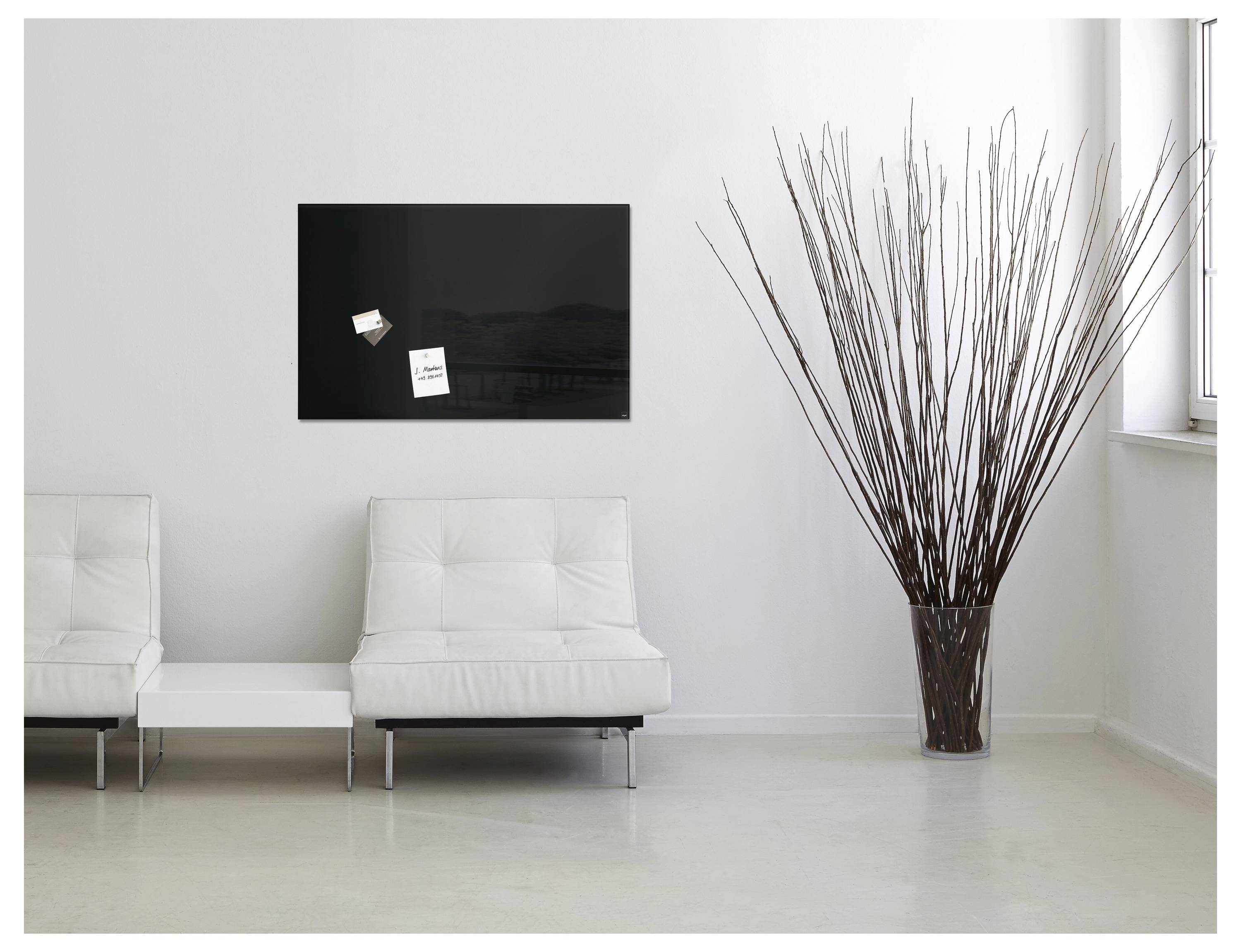 Minimalistisches modernes Wartezimmer mit zwei weißen Stühlen, einem kleinen Beistelltisch, einer großen Vase mit Ästen und einer schwarz angebrachten Wandtafel.