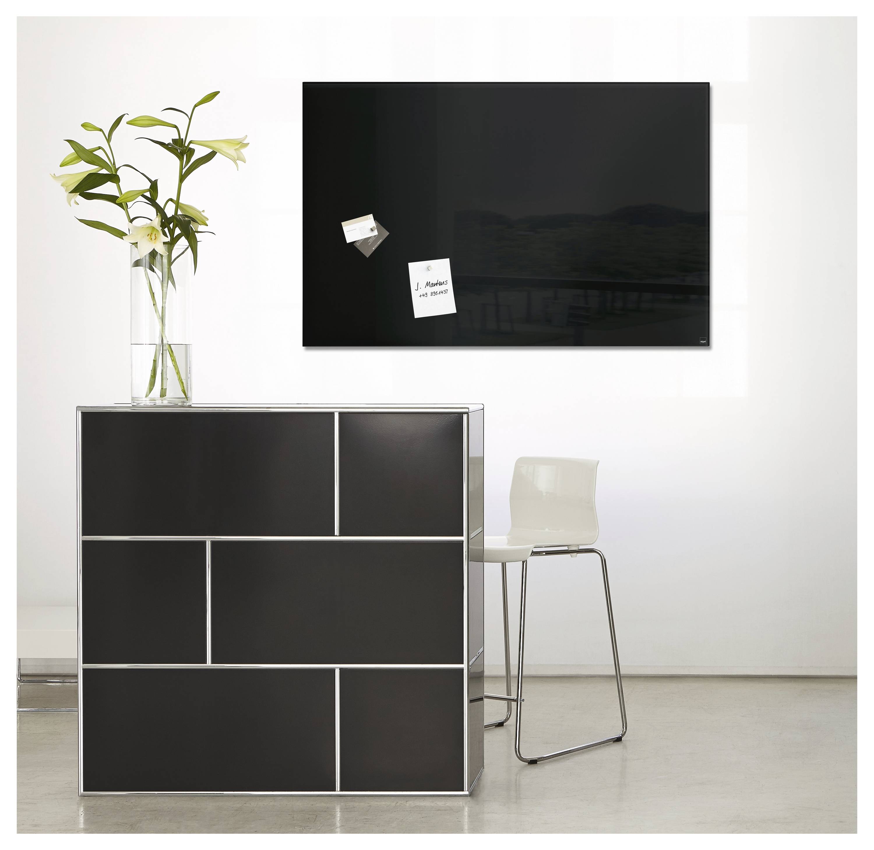 Moderner Raum mit schwarzem modularem Schrank, Vase mit weißen Lilien, wandmontiertem Fernseher und weißem Stuhl auf minimalistischem weißem Boden