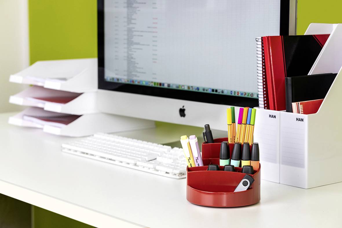 Ein aufgeräumter Schreibtisch mit einem Computerbildschirm, Tastatur, roten Stiftehalter, Stiften, Notizheften und Dokumentablage.
