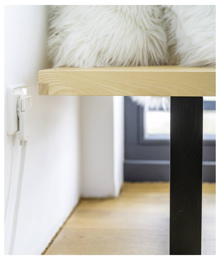 Ein moderner Tisch neben einer Wandsteckdose mit einem eingesteckten Kabel, einem flauschigen Stuhlkissen darüber, und einem Fenster im Hintergrund.
