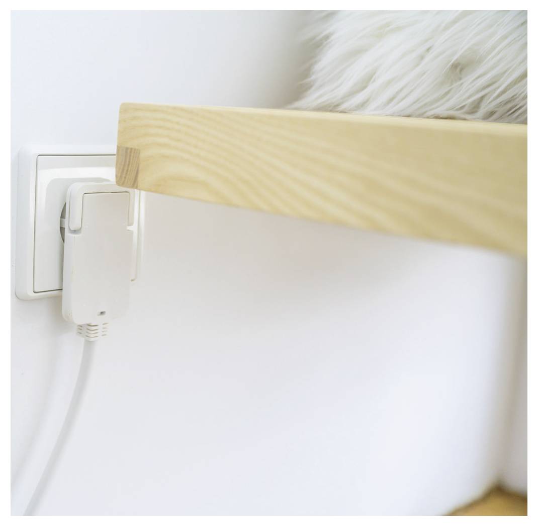 Nahaufnahme einer Wandsteckdose mit angeschlossenem Adapter und Kabel, teilweise verborgen hinter einer Holzmöbelkante neben einer weißen flauschigen Oberfläche.