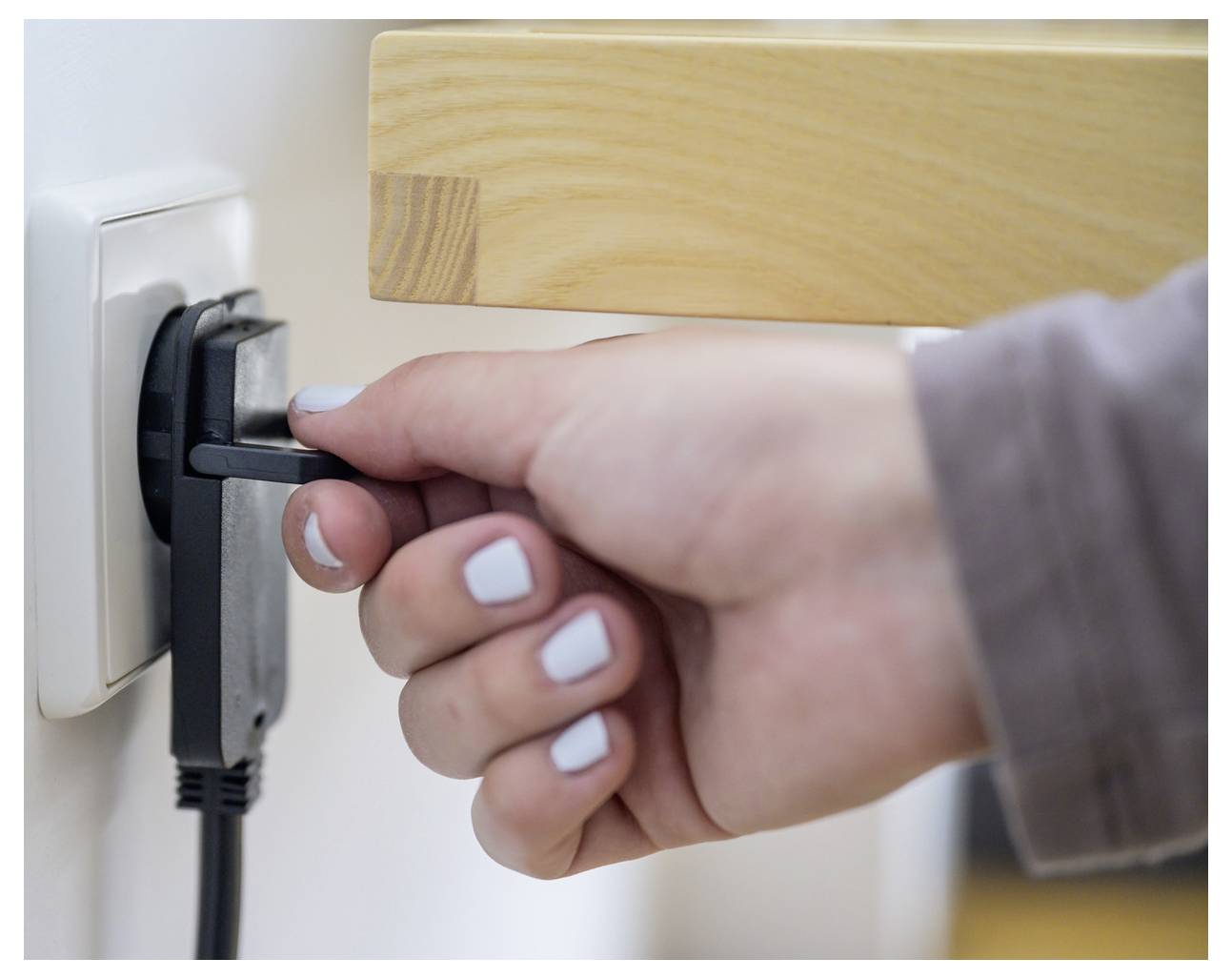 Eine Hand, die ein Stromkabel aus einer Steckdose in der Wand neben einer Holzoberfläche herausgezogen wird.