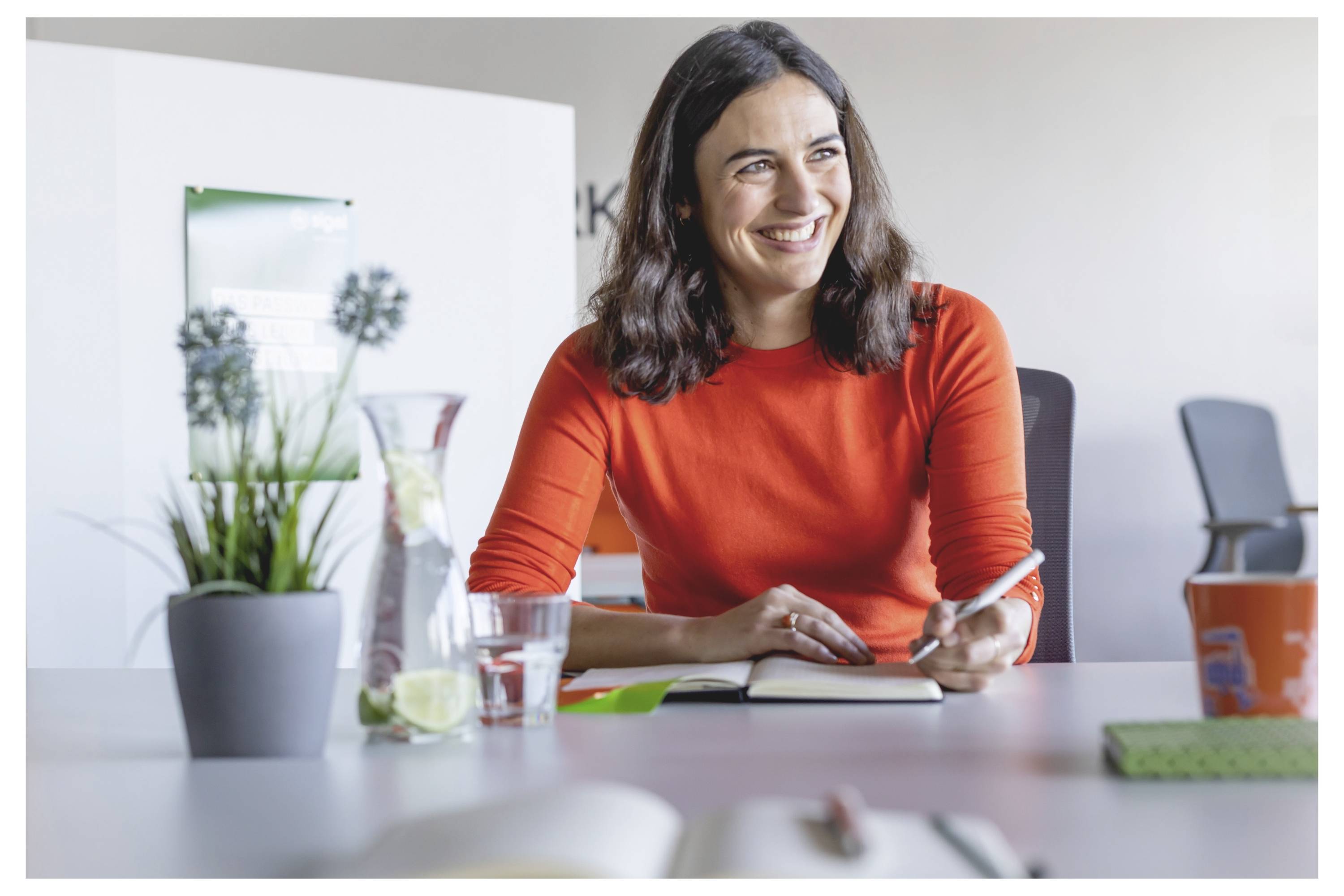 Lächelnde Frau in einem orangefarbenen Pullover sitzt an einem Schreibtisch und hält einen Stift und ein Notizbuch in einem hellen Bürobesprechungsraum.