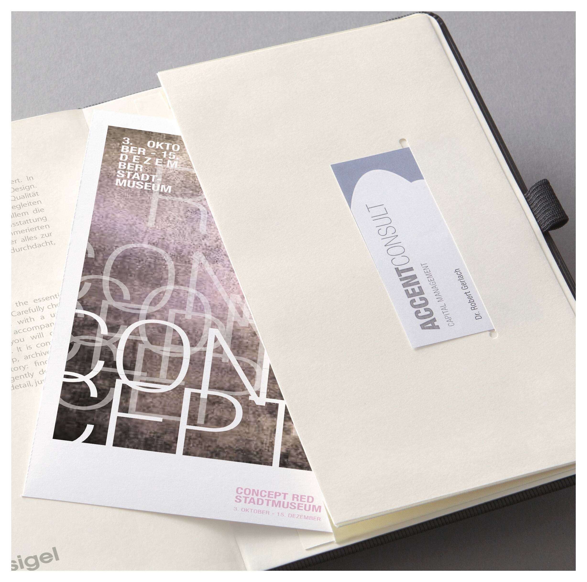 Creme-farbenes Notizbuch mit dem Tag "ACCENTCONSULT" liegt über einer gedruckten Museumsbro schüre auf einem Schreibtisch.
