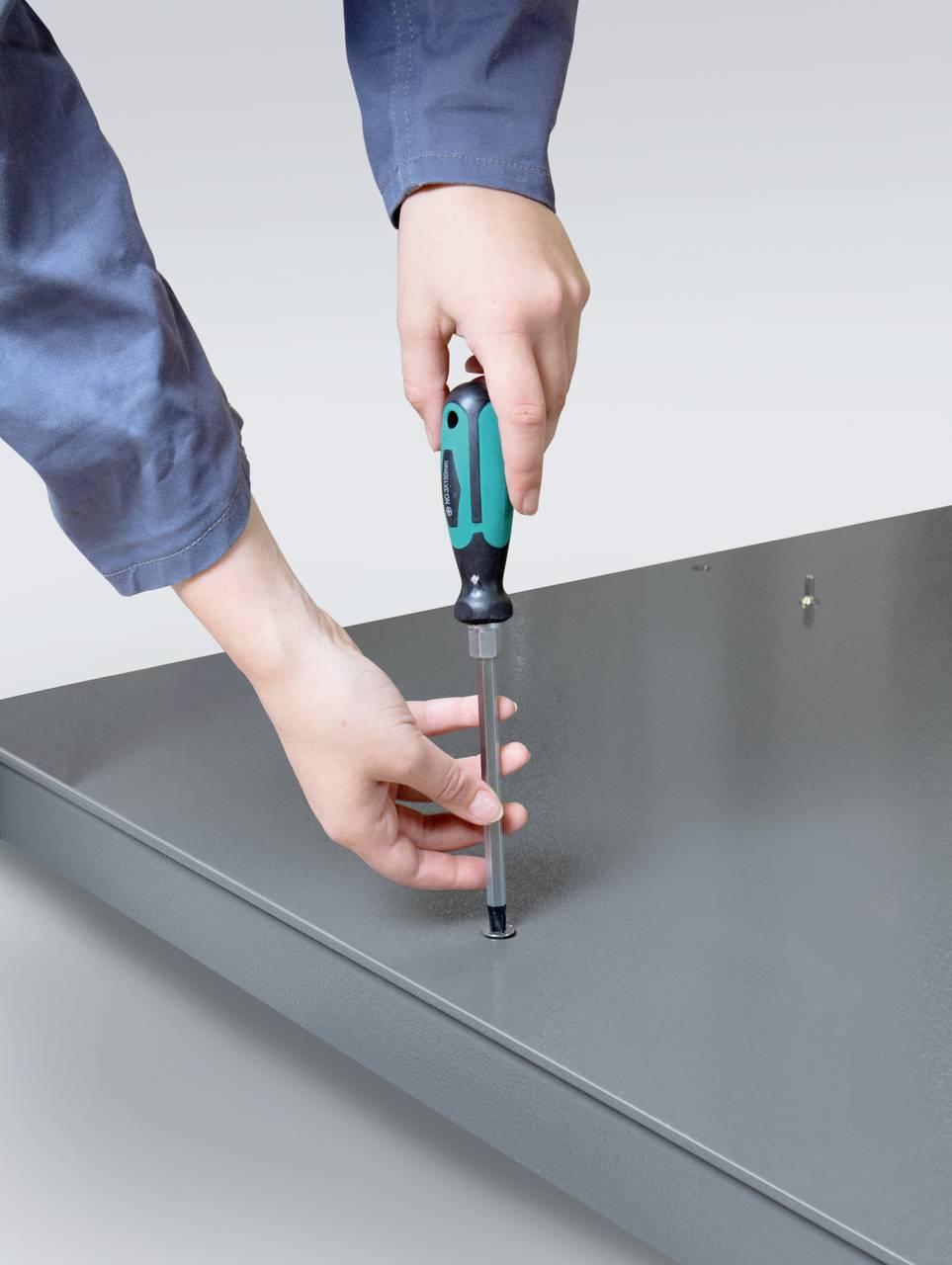 Eine Person schraubt mit einem Schraubendreher eine Schraube in eine graue Metallplatte. Die Hände sind im Fokus der Aufnahme.