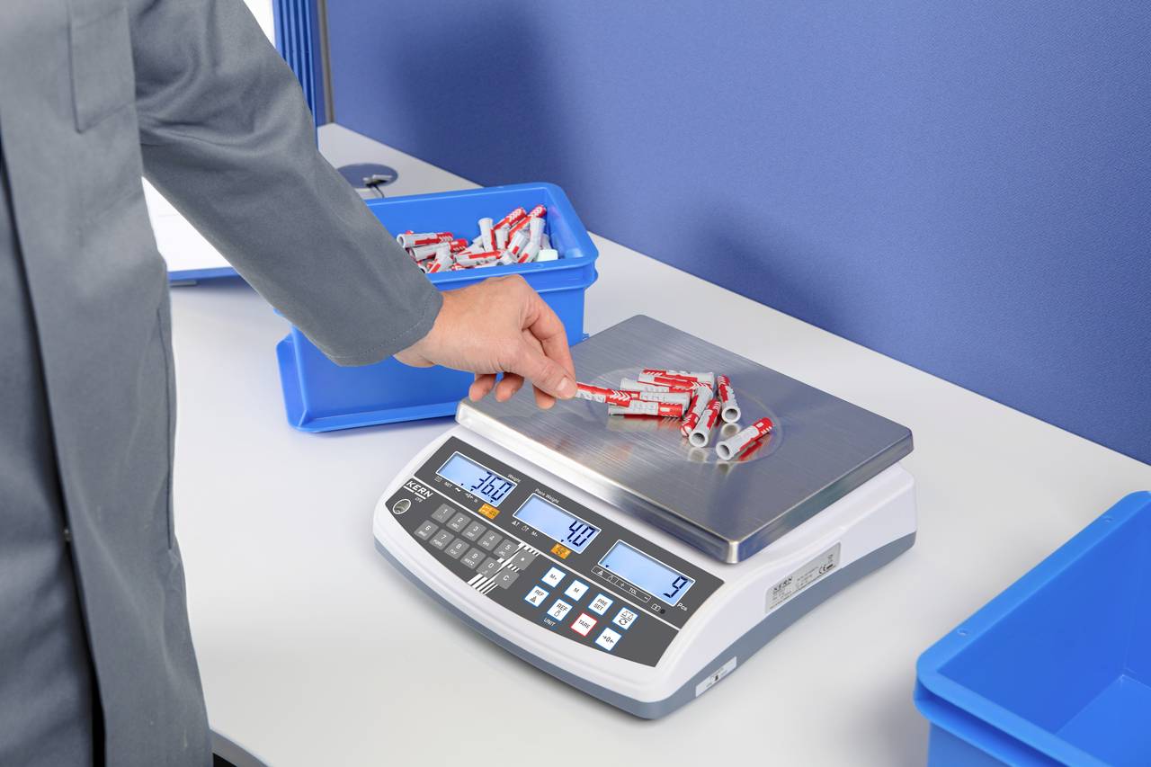 Eine Person wiegt rote elektronische Bauteile auf einer digitalen Waage. Die Waage zeigt ein Gewicht von 240 Gramm.