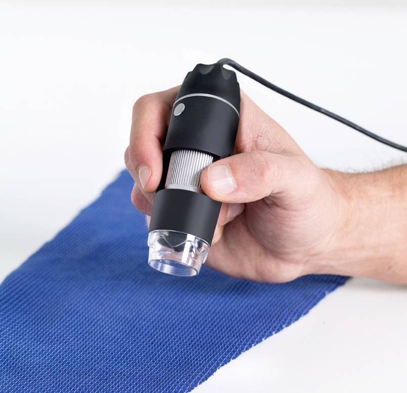 Eine Hand hält ein tragbares Mikroskop über ein blaues Gewebe, um dessen Struktur zu untersuchen.