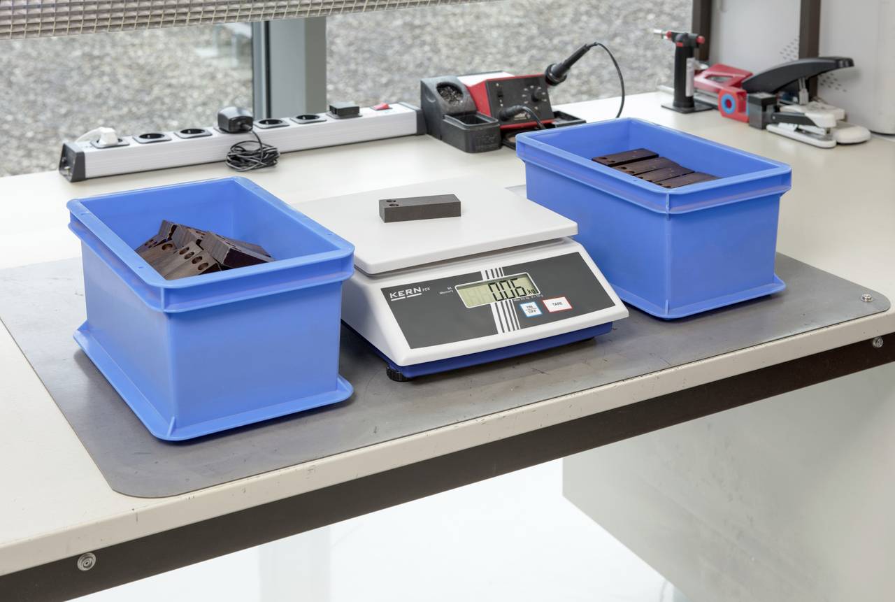 Elektronische Waage auf Tisch mit zwei blauen Behältern. Einer der Behälter enthält Teile, der andere ist leer. Die Anzeige zeigt 0,405 kg.