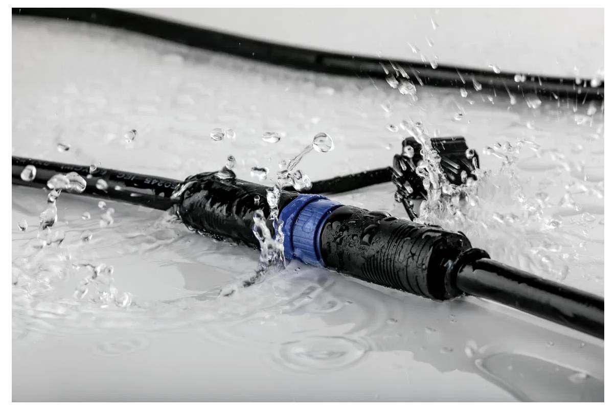 Ein schwarzer Kabelstecker mit einem blauen Ring ist im Wasser zu sehen und demonstriert seine Wasserdichtigkeit. Wassertropfen spritzen um ihn herum.