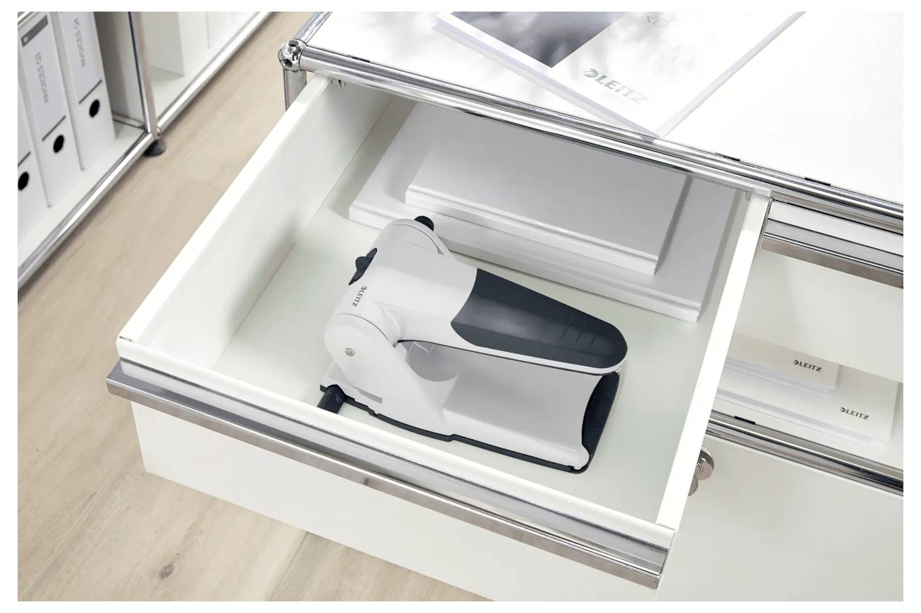 Eine offene Schublade, die ein weißes Elektrogerät auf einem Stapel von Papieren zeigt. Die Schublade ist Teil eines Schranks aus Metall und Holz.