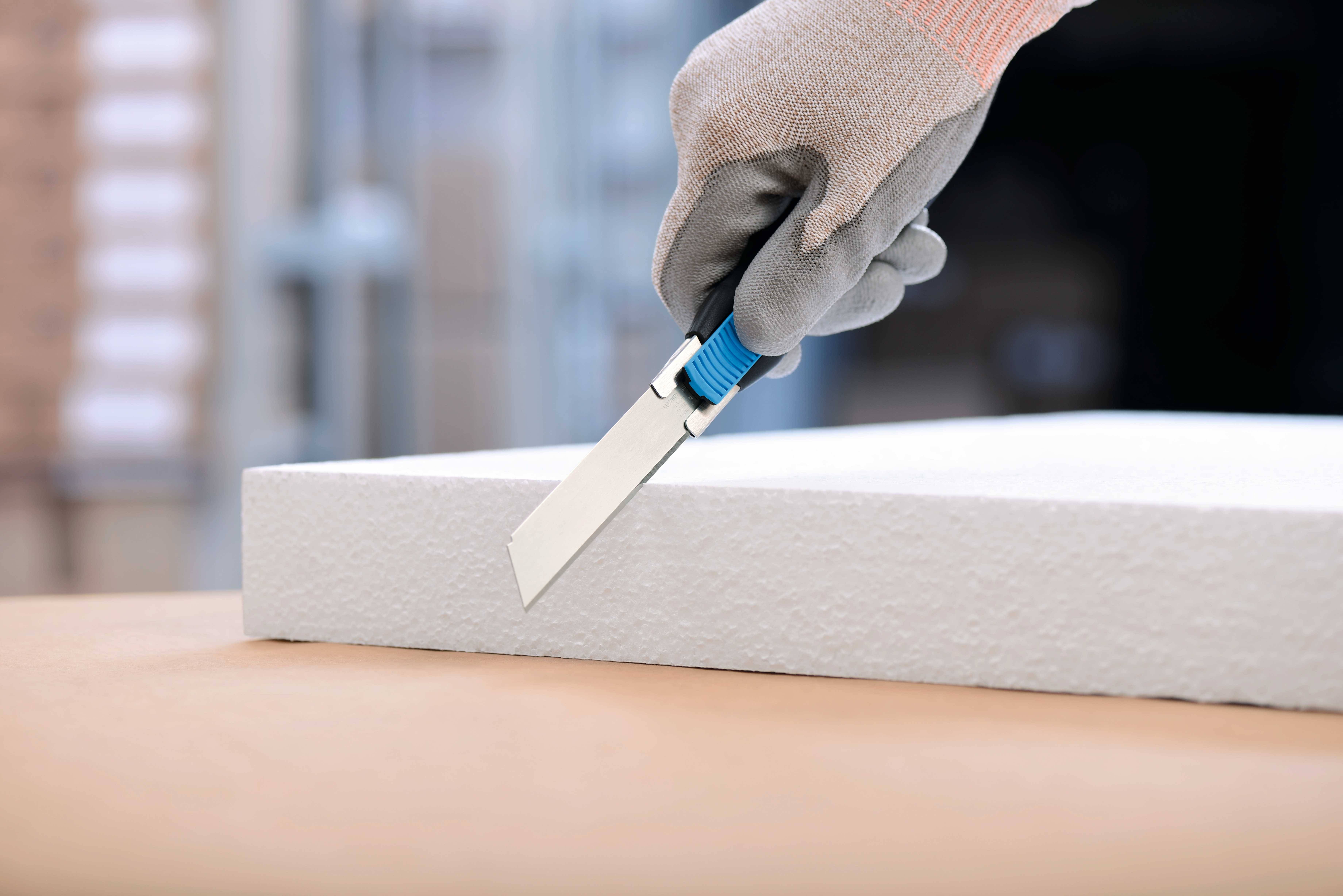Hand mit grauem Handschuh schneidet mit Schneidemesser eine weiße Styroporplatte auf einem Tisch.