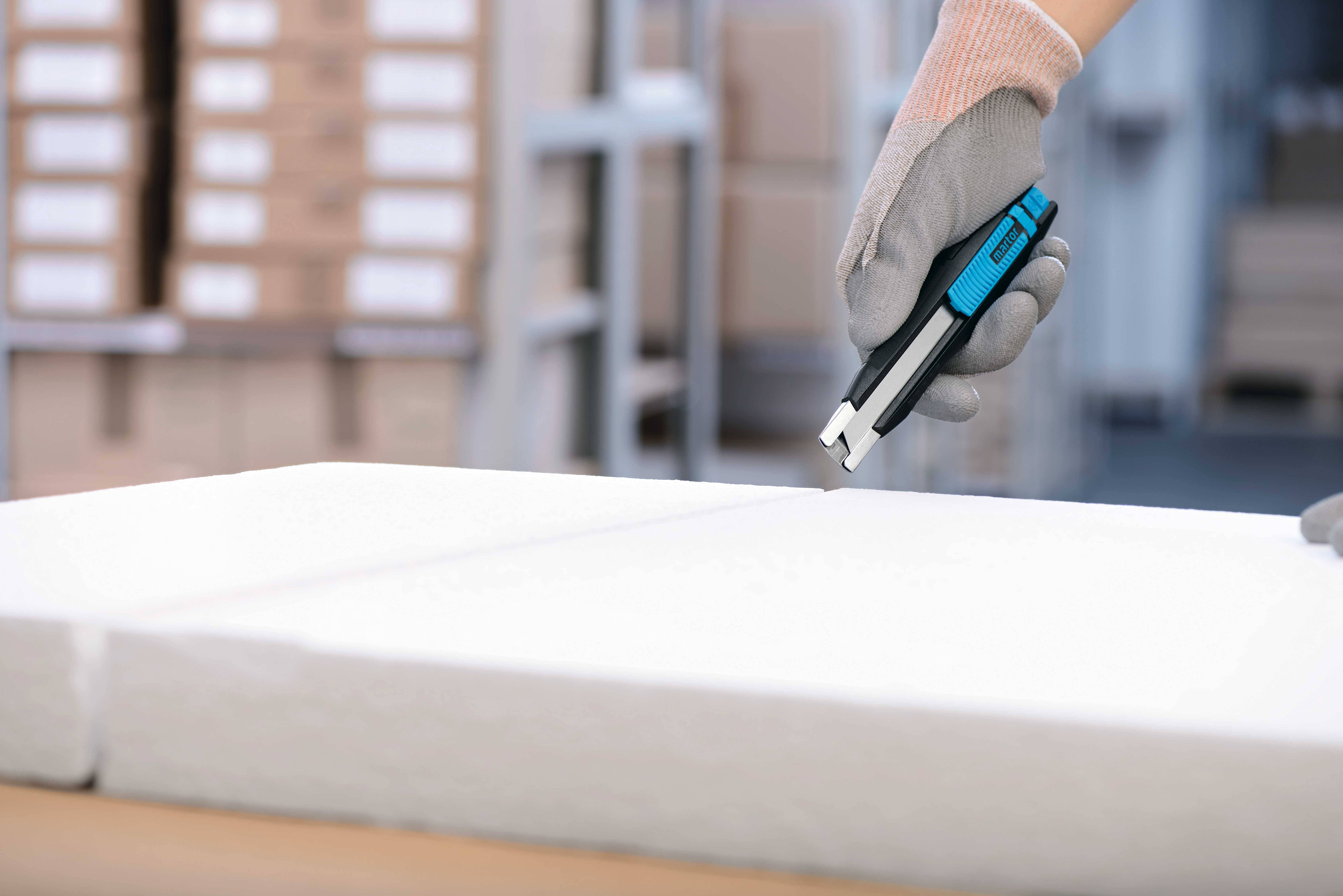 Eine behandschuhte Hand hält ein Teppichmesser und schneidet Styropor in einer Lagerumgebung. Im Hintergrund sind Regale zu sehen.