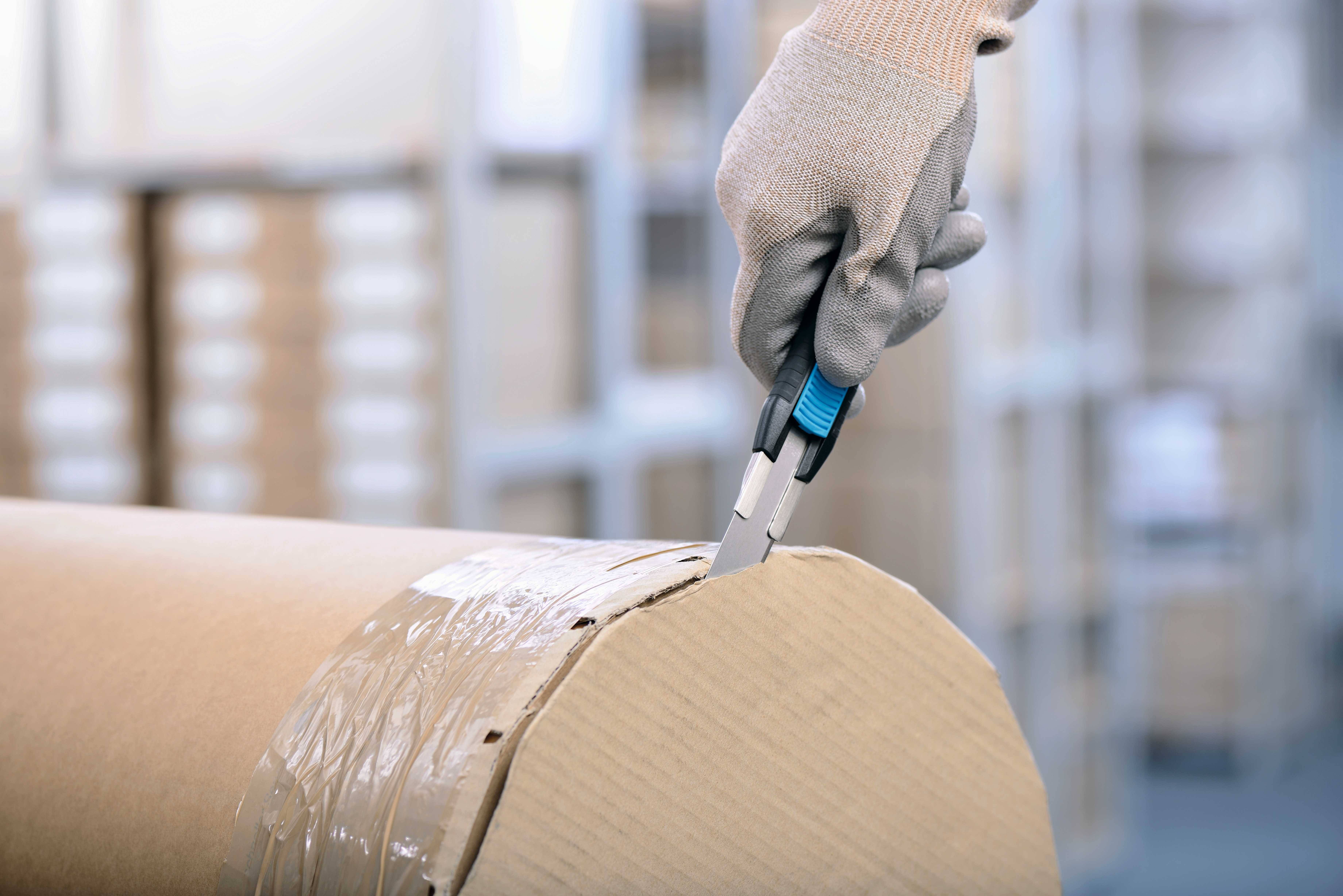 Eine Person schneidet mit einem Kartonmesser eine kartonverpackte Rolle auf. Hintergrund: unscharfe Regale mit Paketen.