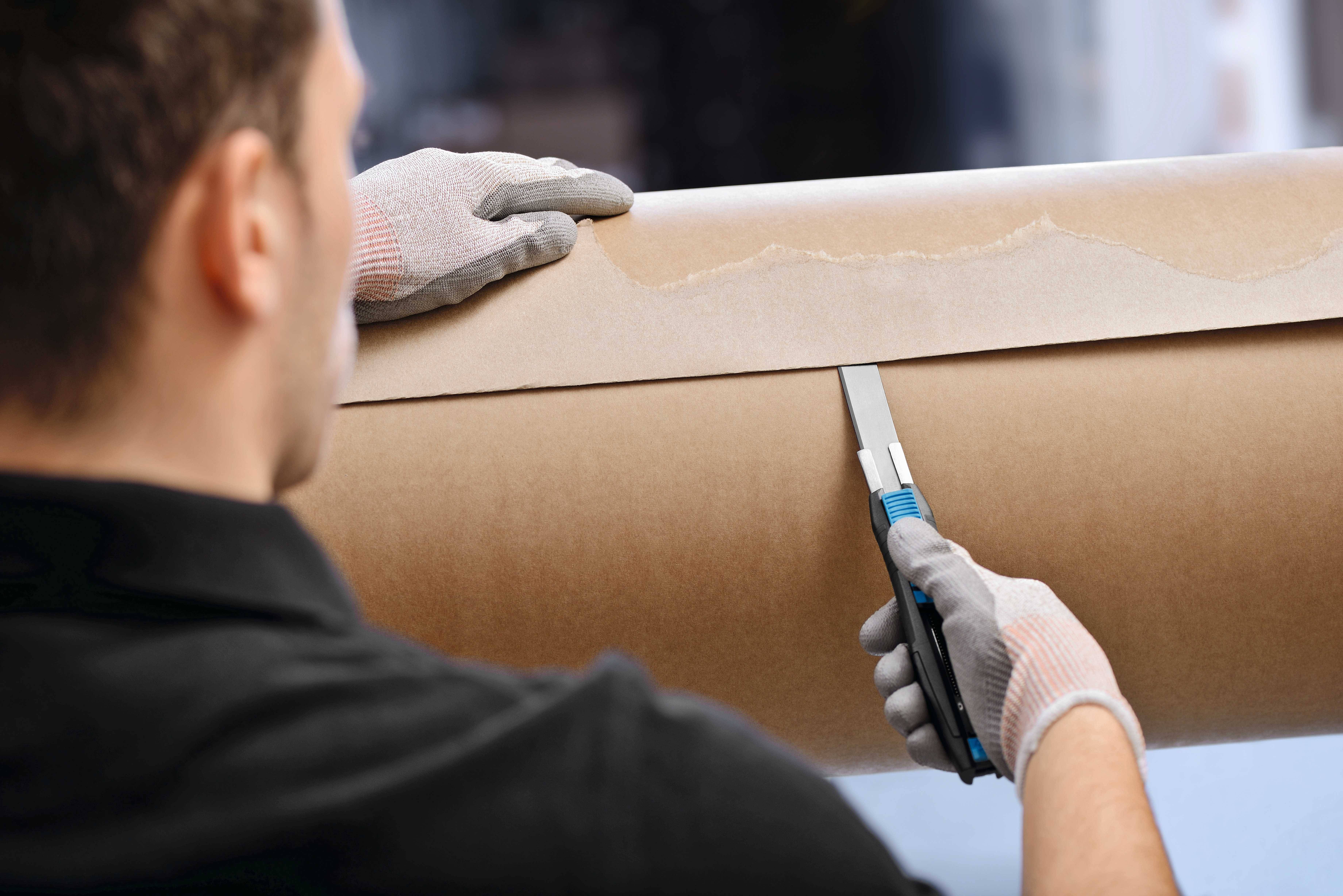 Eine Person schneidet mit einem Teppichmesser ein Blatt Papier auf einer großen Rolle. Sie trägt Schutzhandschuhe.