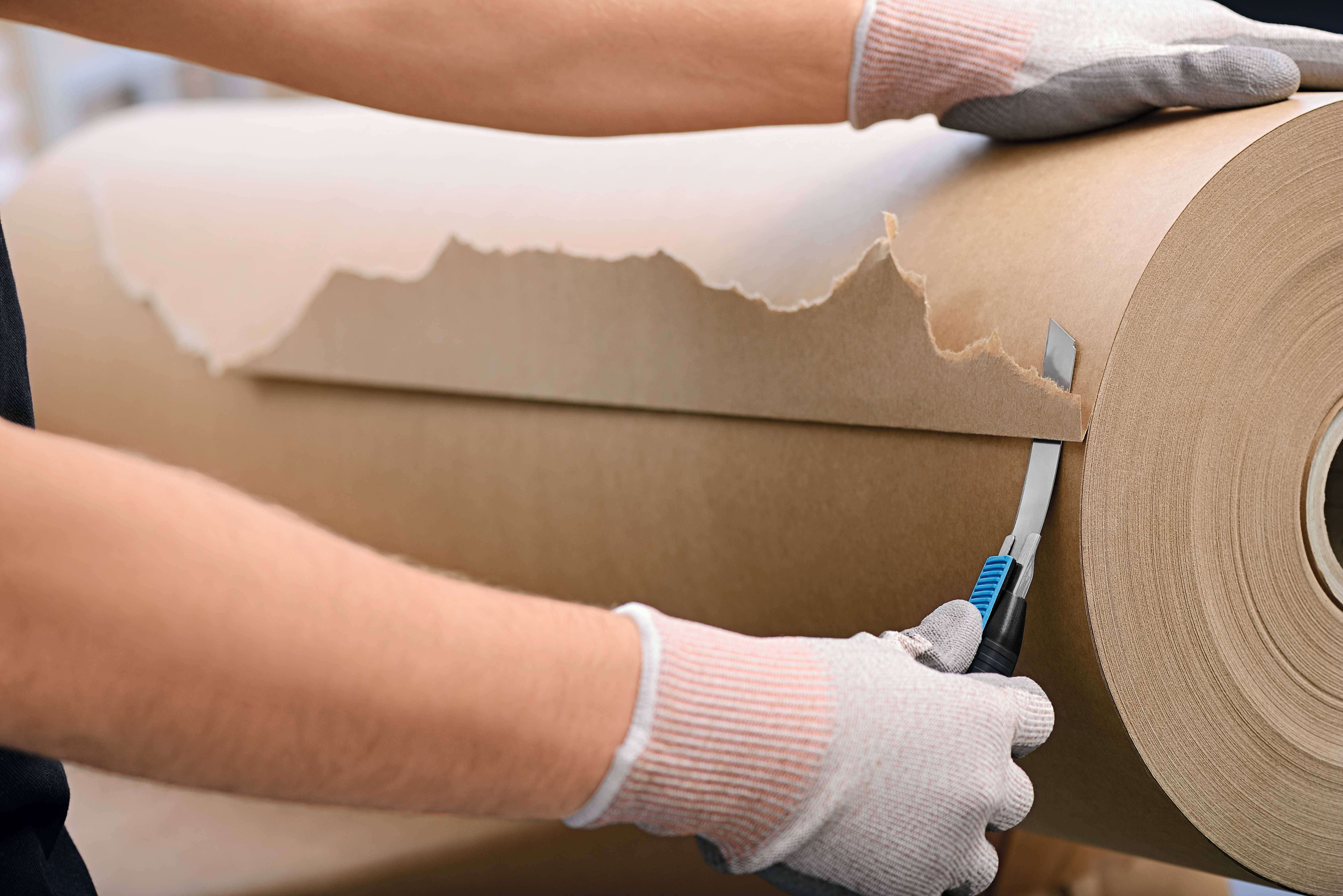Eine Person mit Handschuhen schneidet mit einem Messer eine Rolle dickes Papier ab.