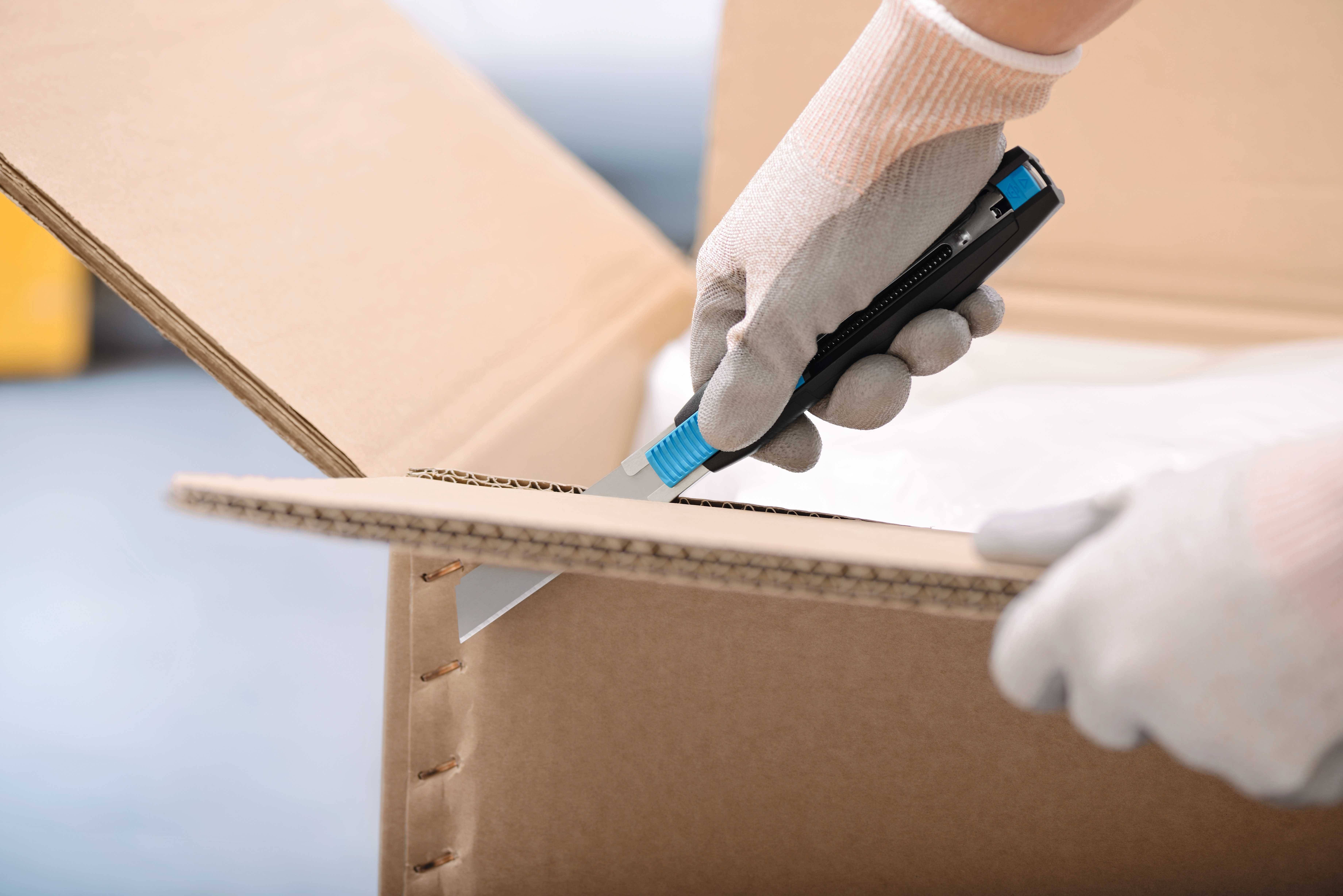 Eine Person mit Handschuhen öffnet mit einem Cuttermesser einen Pappkarton.