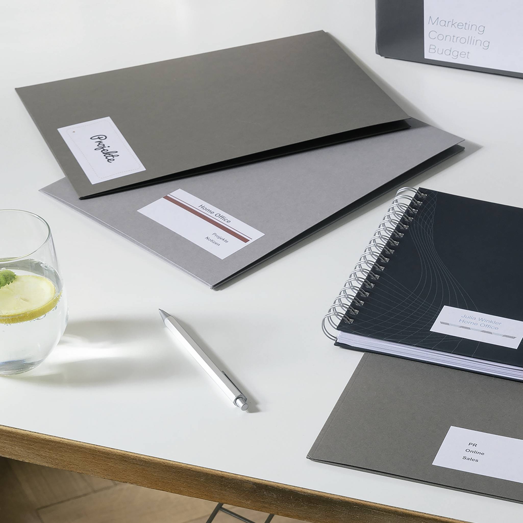 Ein Tisch mit Unterlagen, Notizbüchern und einem Glas Wasser. Auf einem Ordner steht 'Marketing Controlling Budget'. Ein Kugelschreiber liegt neben den Unterlagen.