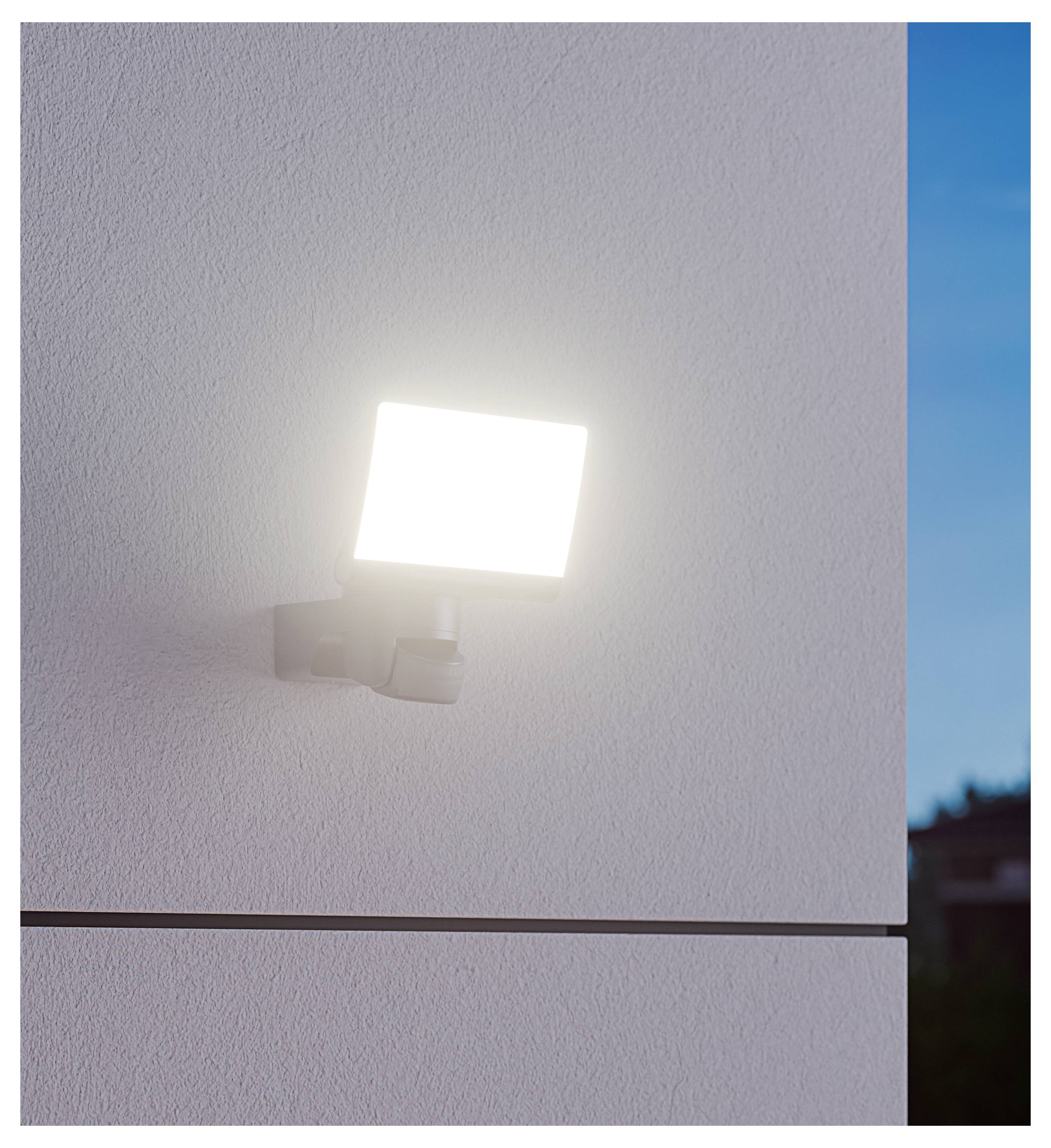 Helle an der Wand montierte Außenleuchte, die eine strukturierte weiße Gebäudefassade in der Dämmerung beleuchtet.