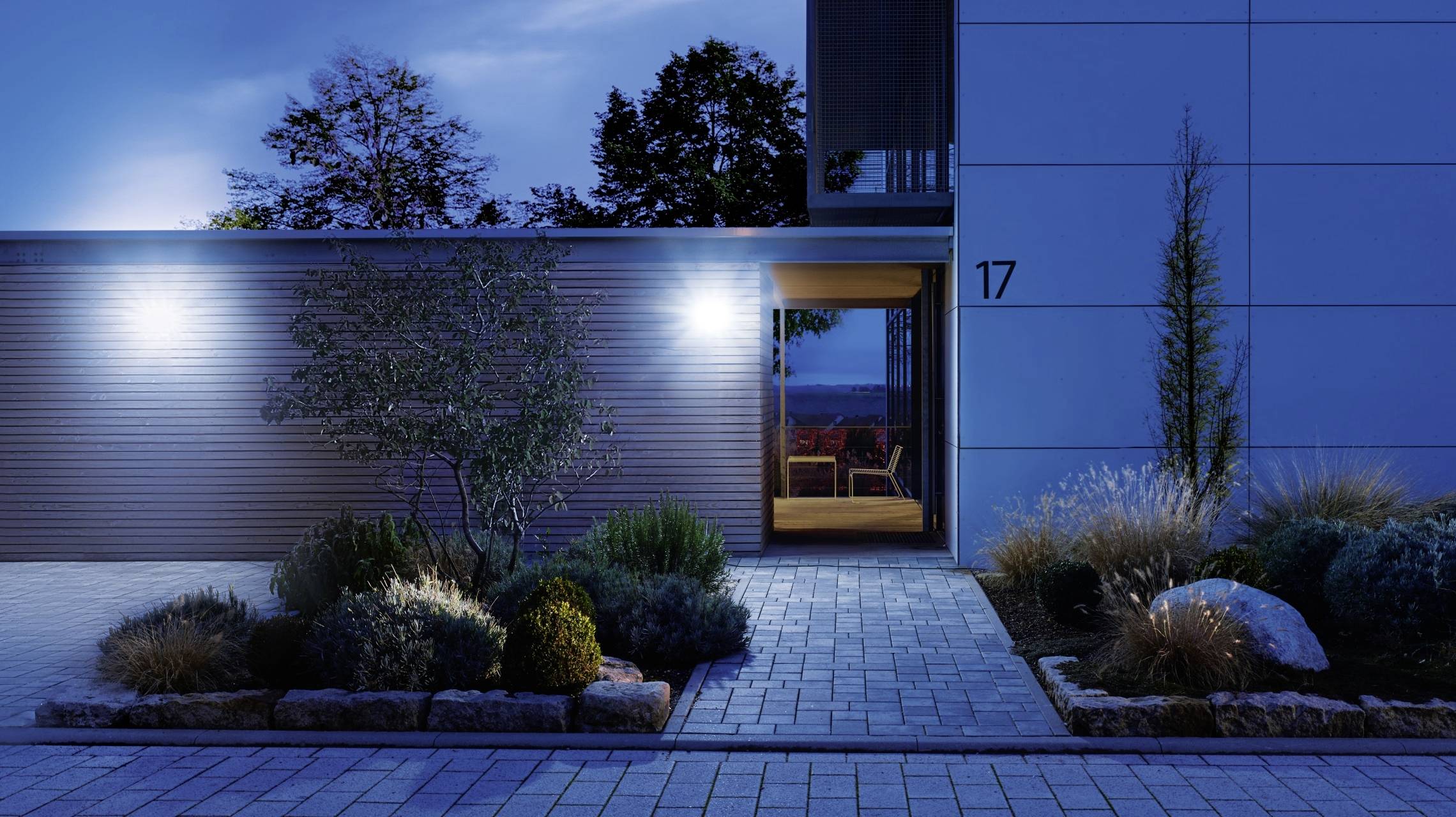 Eingangsbereich eines modernen Hauses bei Nacht. Beleuchteter Weg mit Pflanzen, Bäume im Hintergrund. Hausnummer '17' sichtbar.