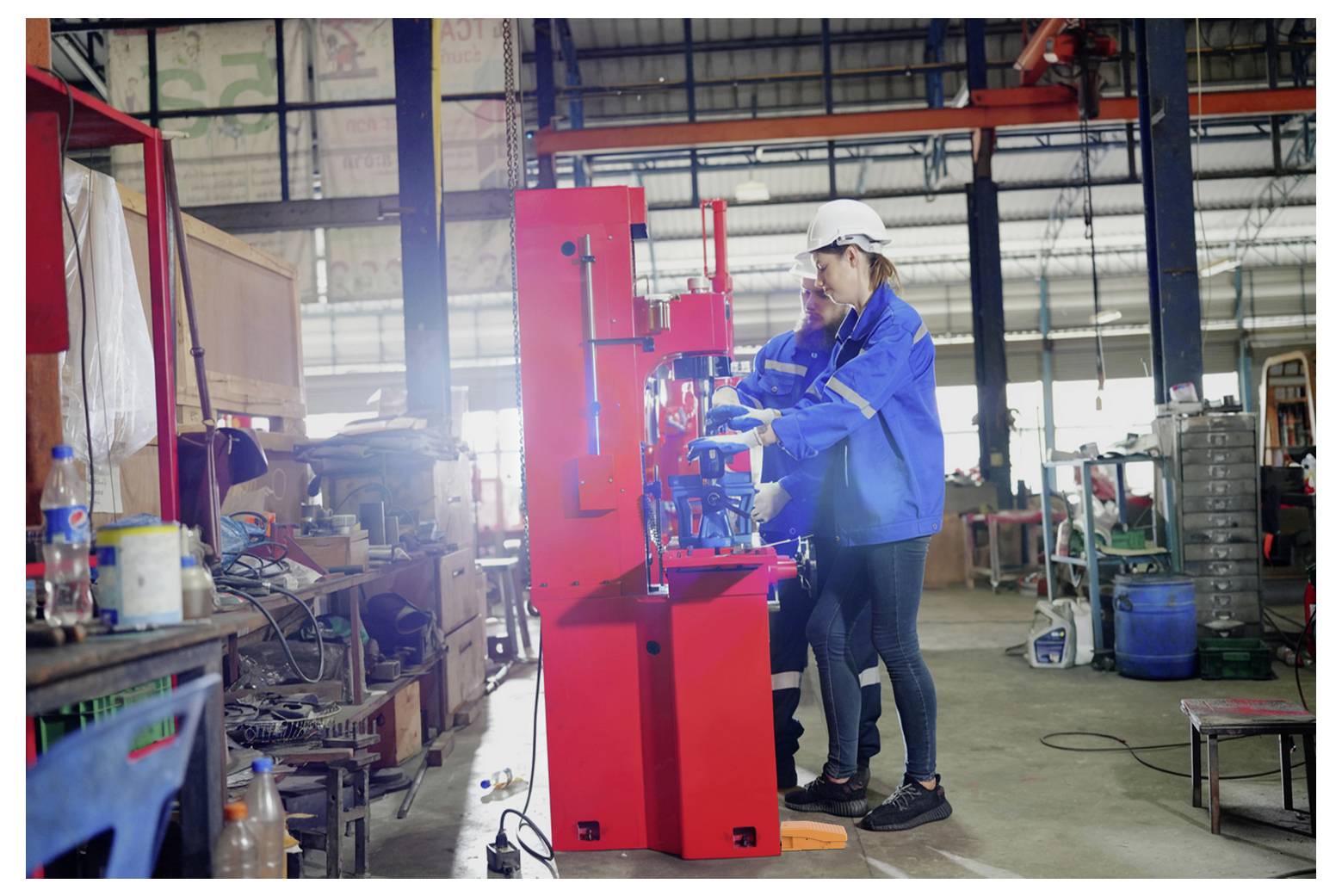 Zwei Fabrikarbeiter in Schutzausrüstung bedienen eine leuchtend rote Industriemaschine in einer Werkstatt oder einem Lagerhaus.