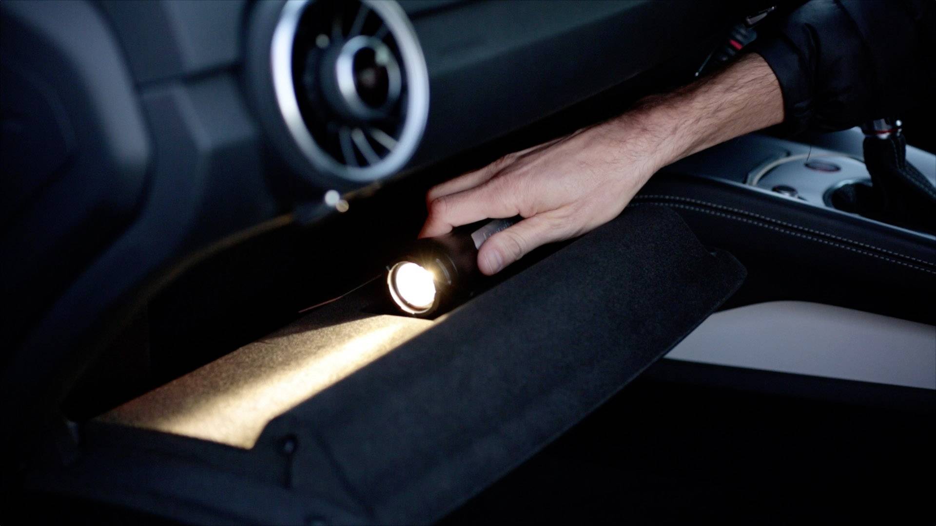 Eine Hand hält eine Taschenlampe und leuchtet in das geöffnete Handschuhfach eines Autos.