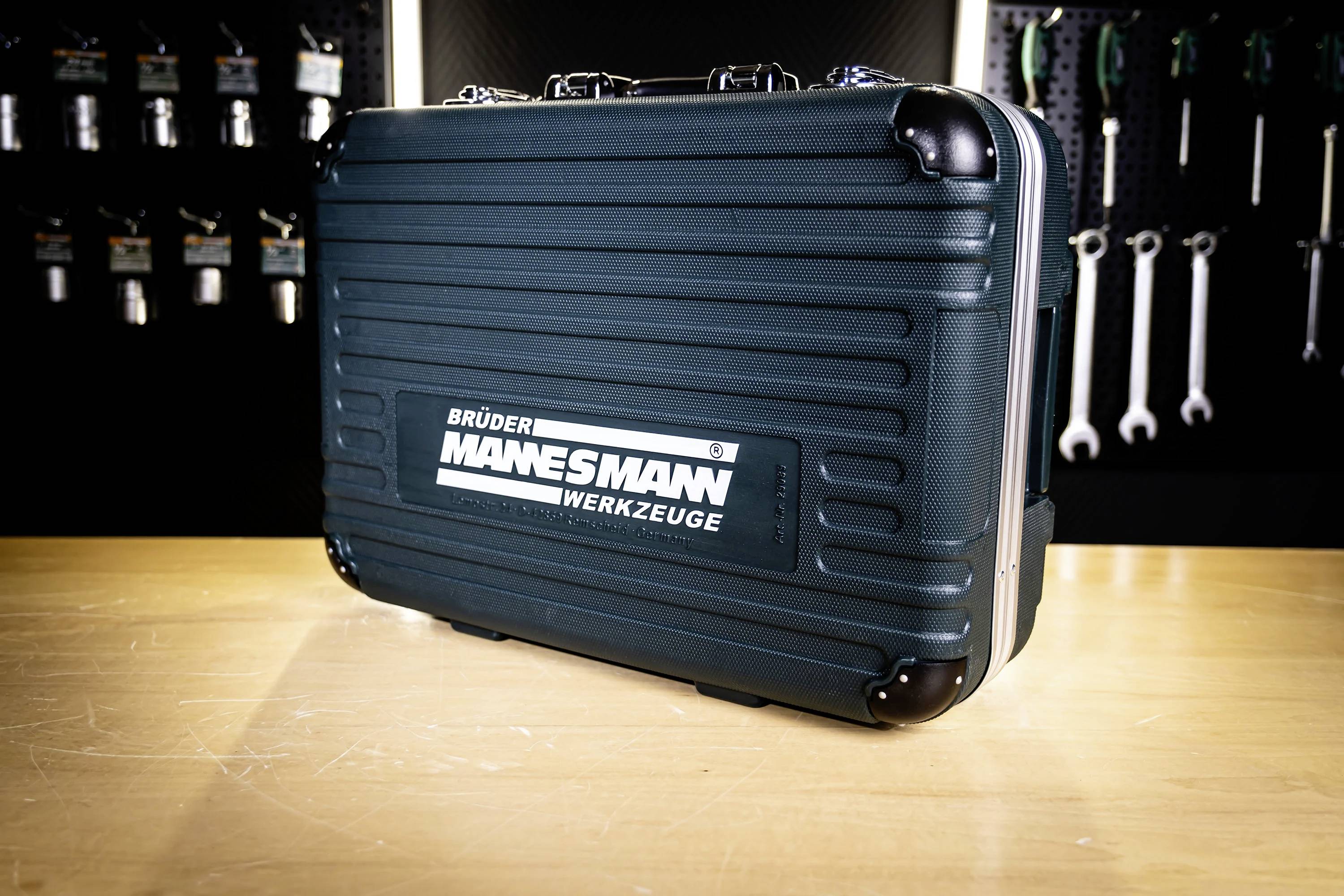 Ein schwarzer Werkzeugkoffer mit der Beschriftung 'Brüder Mannesmann Werkzeuge' auf einem Holztisch. Der Hintergrund zeigt an der Wand hängende Werkzeuge.