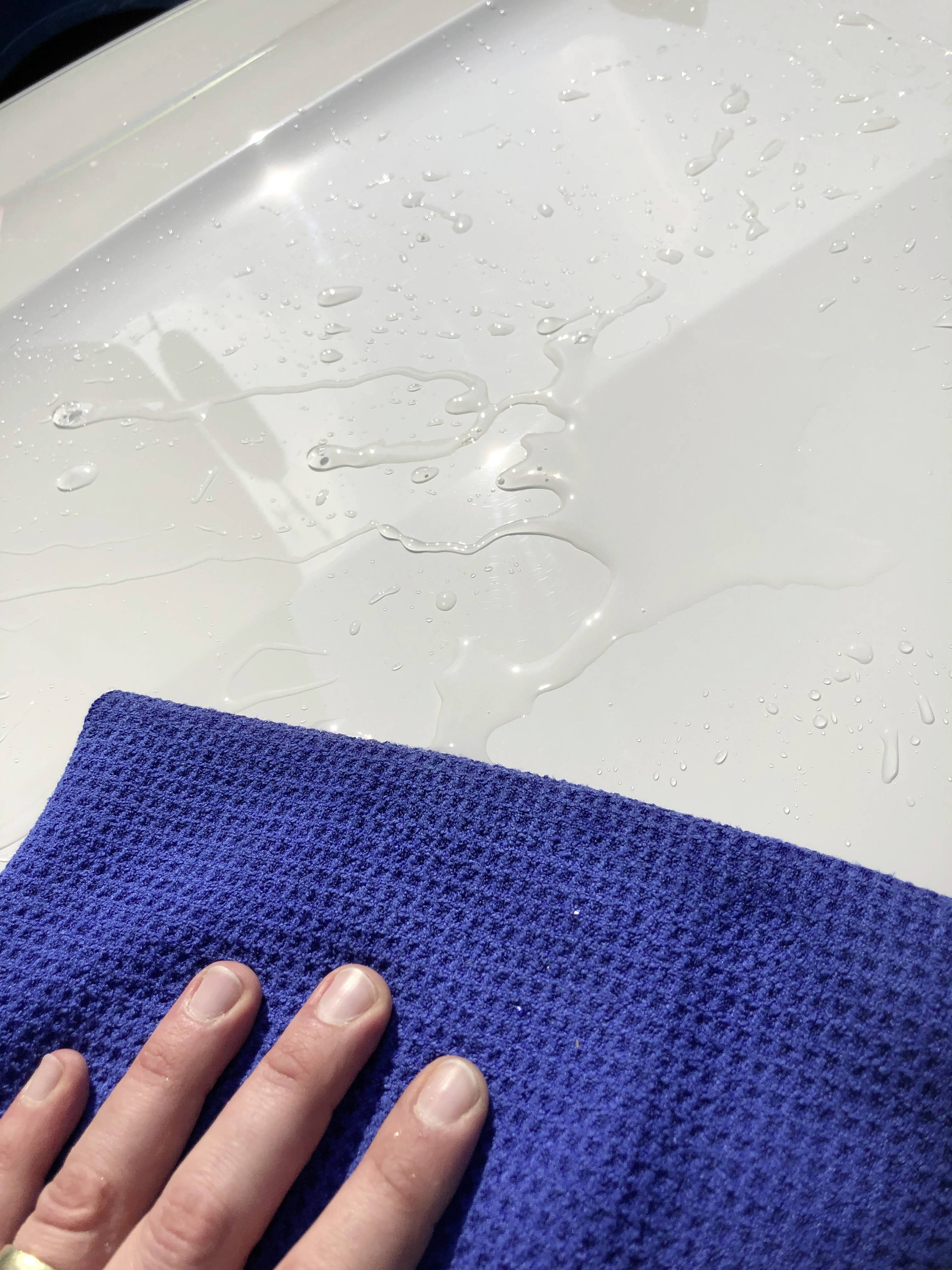 Eine Hand mit einem blauen Tuch wischt Wasser von einer glänzenden weißen Oberfläche. Reflektiertes Licht zeigt Sauberkeit an.