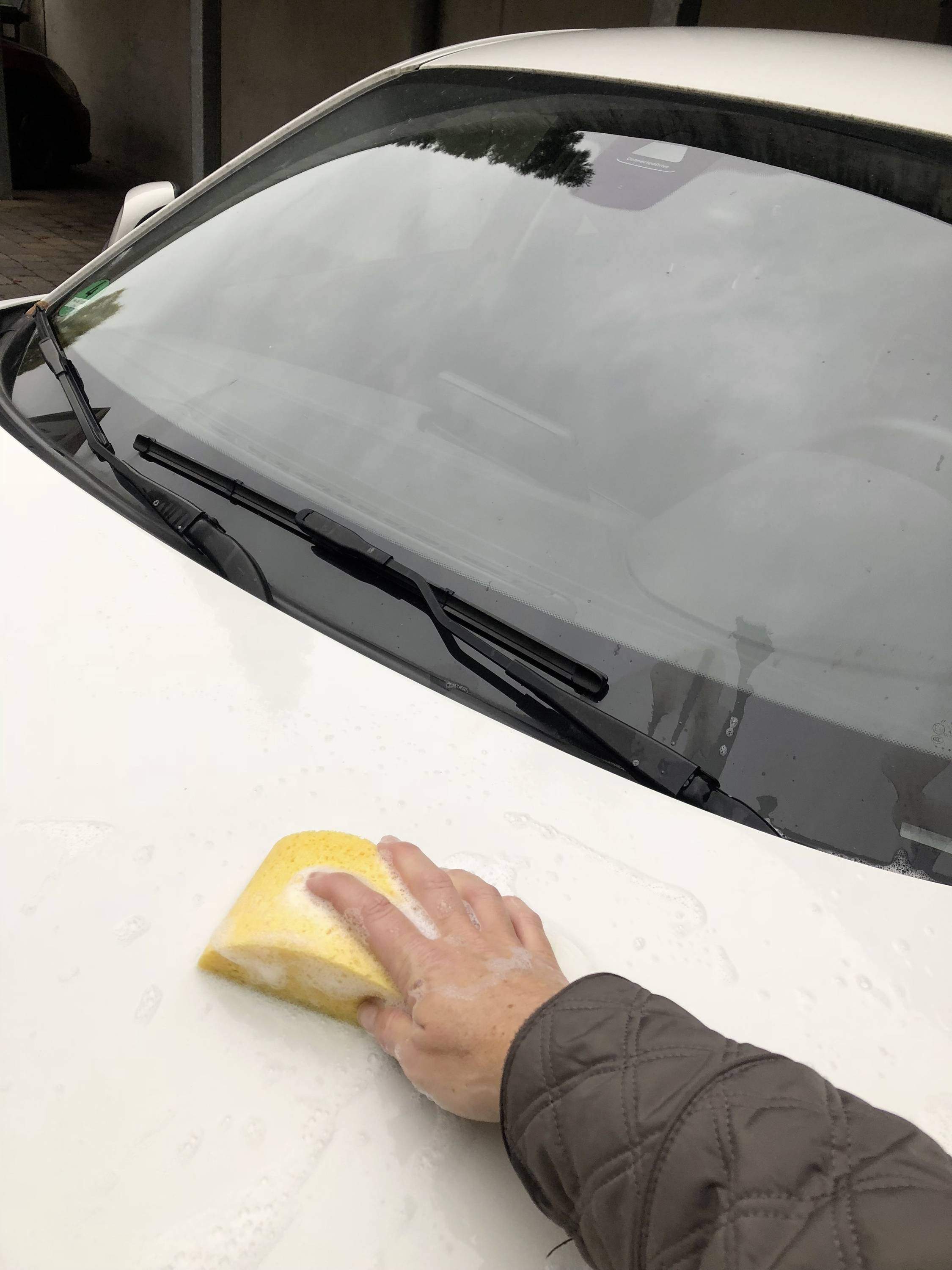 Eine Person reinigt die Motorhaube eines weißen Autos mit einem gelben Schwamm. Das Auto steht in einem Hof.
