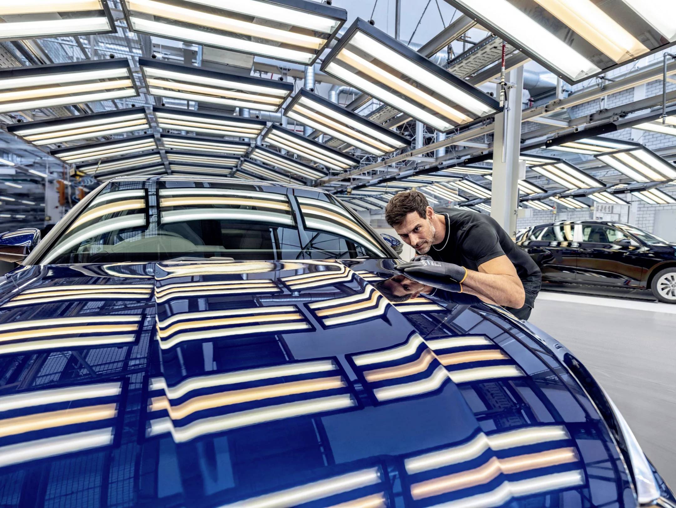 Ein Mann inspiziert sorgfältig die polierte Motorhaube eines blauen Autos in einer hell beleuchteten Fabrikhalle.