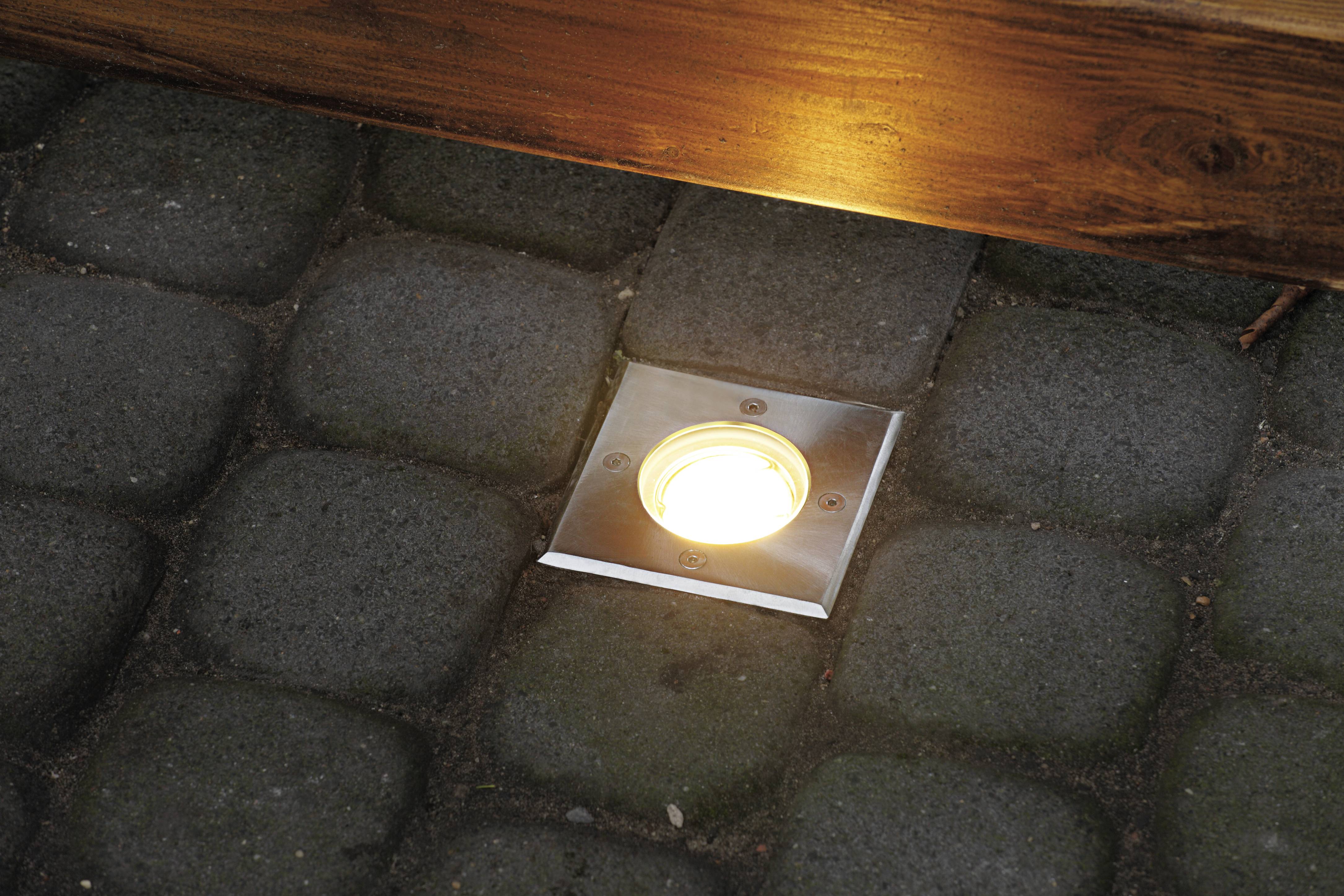 Ein eingelassenes Bodenlicht in einem Pflasterstein-Weg leuchtet hell und beleuchtet den Bereich vor einer Holzkante.